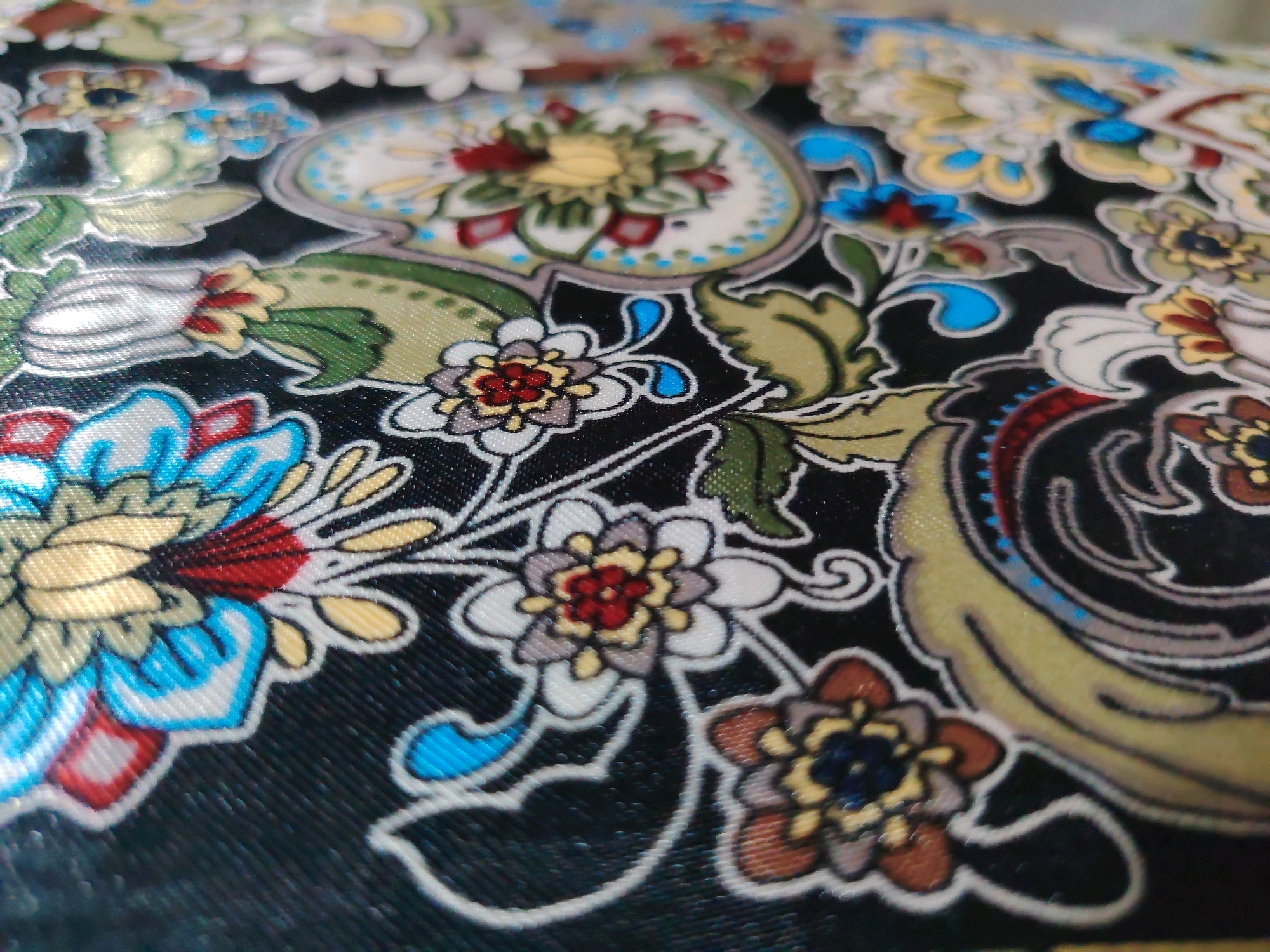 The image shows a close-up, slightly blurry view of a black fabric with a colorful floral pattern. The pattern features stylized flowers and paisley-like motifs in shades of yellow, blue, red, green, and white, outlined in a thin white line. The fabric appears to have a slight sheen, suggesting it might be silk or satin. The perspective is from a low angle, looking across the surface of the fabric. There is no discernible location within Davao City, Philippines.