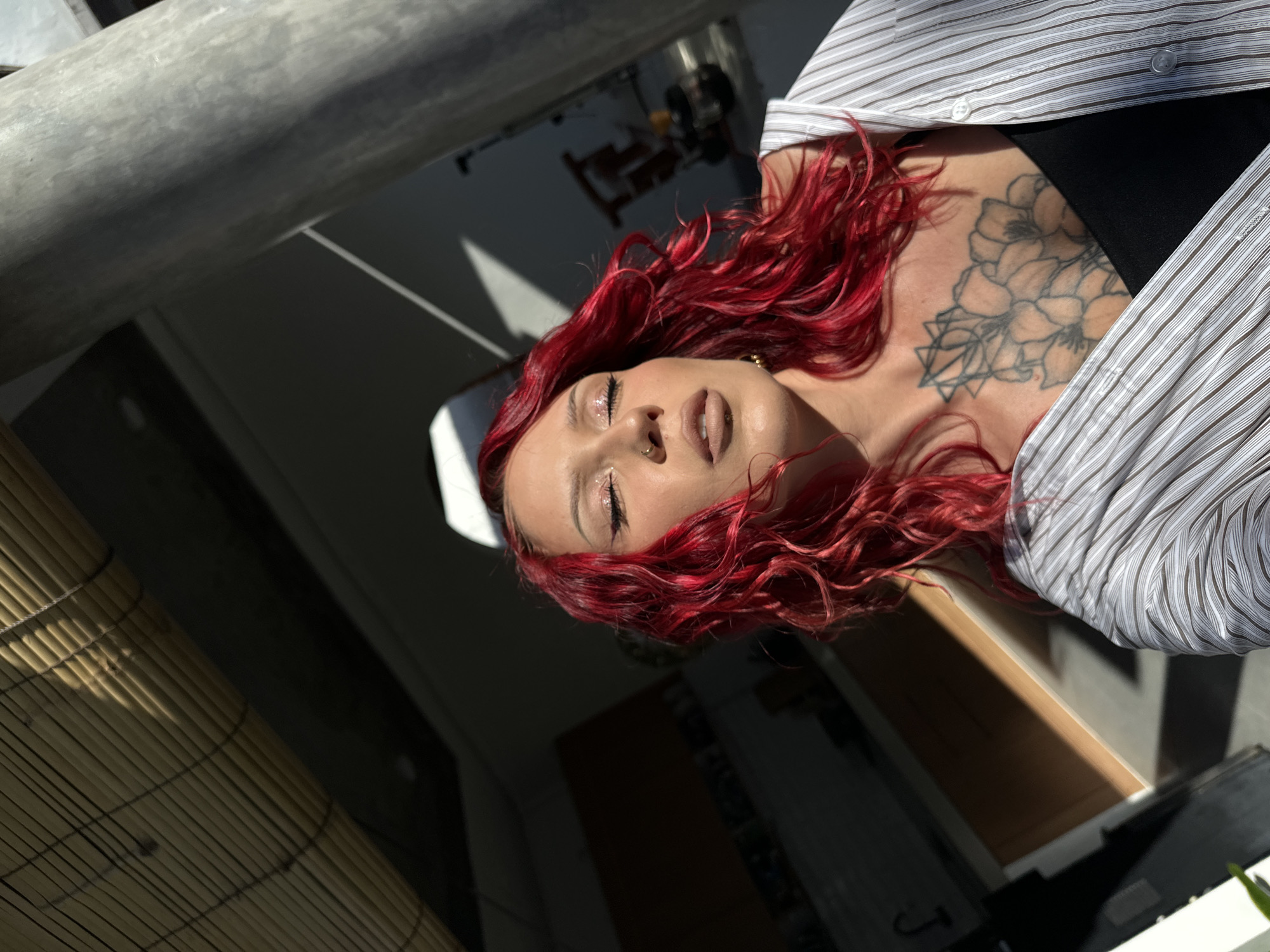 An artistic, horizontally oriented close-up photograph captures a woman with striking, wavy red hair. She is seen from an elevated perspective, lying down or tilted, with her head and upper torso angled across the frame. Her eyes are closed, adorned with glittery eye makeup, and her lips are slightly parted, revealing a nose piercing. She wears a light-colored, striped button-up shirt over a black top, revealing a prominent, uncolored floral tattoo with geometric elements on her upper chest and shoulder area.
Strong, direct sunlight illuminates her face and hair, creating sharp highlights and shadows, indicating a sunny daytime setting, likely morning or late afternoon. A large, grey cylindrical object, possibly a metal beam or pole, cuts diagonally across the top left of the frame. In the bottom left, a bamboo blind or mat is visible, partially illuminated by the sun, suggesting an indoor or semi-outdoor space. The background behind the woman is dark and out of focus, showing indistinct architectural elements. The presence of bamboo and bright sunlight aligns with the specified location context of North Kuta, Indonesia. No visible text is present.