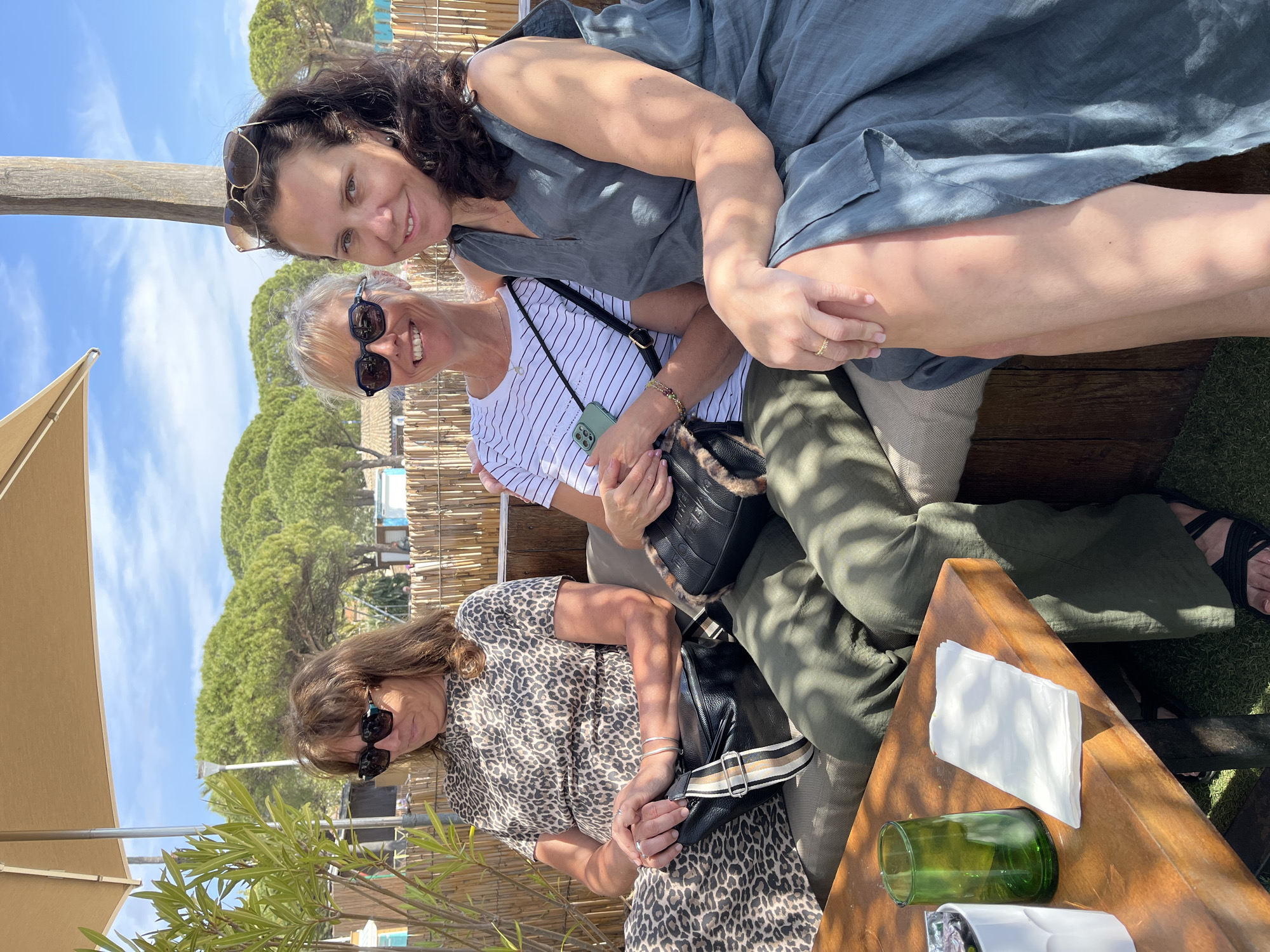 Three women are seated together outdoors in Sainte-Maxime, France, at a café. One woman, with dark curly hair, is wearing a gray dress and sunglasses on her head. The second woman has short gray hair, sunglasses, and a striped shirt; she is holding a phone and a black purse. The third woman wears a leopard-print dress and sunglasses, with a black purse at her side. They are sitting on a bench or lounge seating with cushions. A wooden table is nearby with napkins and glasses. The setting includes a bamboo fence, trees, and a blue sky with white clouds. A sunshade is partially visible.