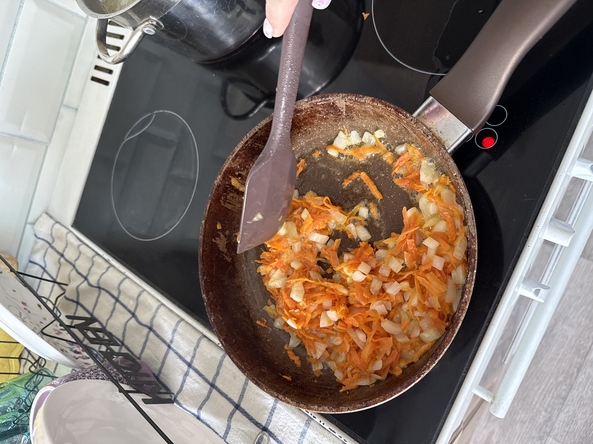 The image shows a close-up, eye-level view of a non-stick frying pan on a black electric stovetop. The pan contains chopped onions and shredded carrots that are being sauteed in oil. A brown spatula is being used to stir the vegetables, and a hand with light-colored fingernails is visible to the right, holding the spatula. To the left of the pan, a white and blue checkered dishtowel is laid out on a kitchen counter, next to a dish rack holding plates. In the background, white subway tiles form the backsplash of the kitchen wall. A stainless steel pot with a handle is situated on the stovetop to the right of the frying pan. The stovetop has illuminated red circles, indicating that burners are active. This scene appears to be taking place in a kitchen. The specific location of Stary Oskol, Russia, cannot be determined from the visual information provided in the image.