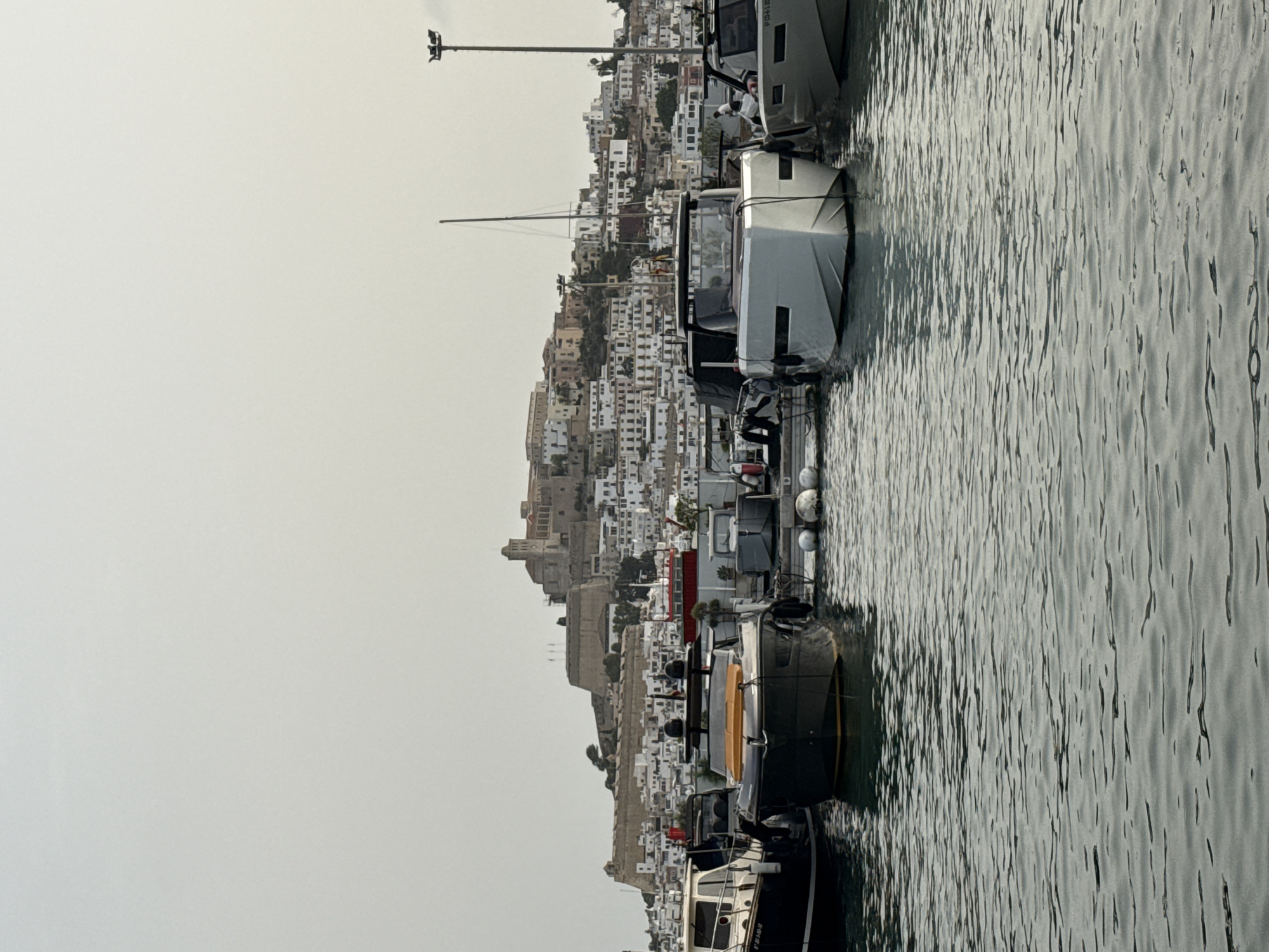 The image depicts a view of the port of Eivissa, Spain, with the historic Dalt Vila citadel prominently visible on the hill in the background. Several yachts and boats are moored in the foreground, bobbing gently on the rippling water. The sky is overcast, casting a soft, diffused light over the scene. The architecture of Dalt Vila, a UNESCO World Heritage site, features a mix of fortified walls and white-washed buildings cascading down the hillside. A few streetlights are visible on poles in the marina area. The overall impression is of a calm, European coastal town, possibly in the late afternoon or early morning given the subdued lighting.