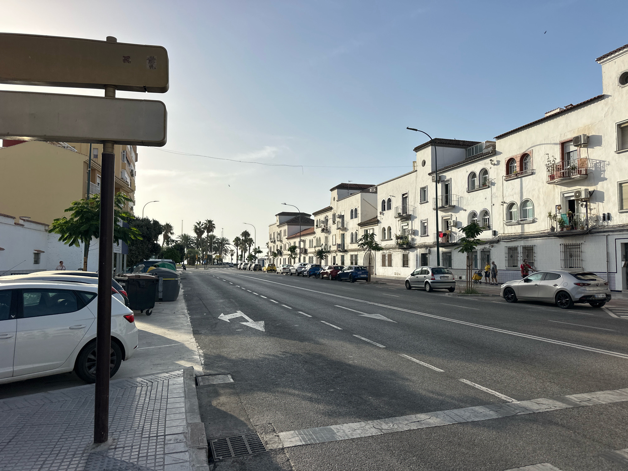The image depicts a street scene in Málaga, Spain, likely along the coast given the presence of palm trees and the style of architecture. A white car is parked in the foreground on the left, partially obscuring a sidewalk with a textured pattern. A dark pole stands near the sidewalk. Across the street, a row of white buildings with Spanish architectural elements, such as arched windows and balconies, lines the road. Several cars are parked along the street, and a few people are visible walking on the sidewalk in front of the buildings. The road has clear lane markings, including directional arrows. In the background, more palm trees and buildings are visible under a clear blue sky. Several large green and brown trash bins are situated near the sidewalk on the right side of the street. The scene appears to be during daylight hours, with sunlight casting shadows on the road.