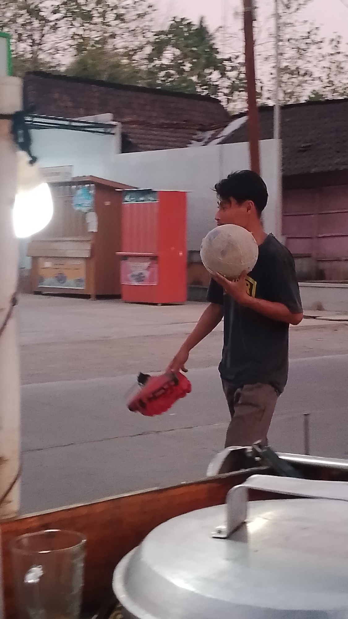 A young man is walking, holding a soccer ball in his right hand and a red shoe in his left hand. He is wearing a dark t-shirt and light brown pants. He is in an outdoor environment, possibly a street or a public area, with buildings and trees in the background. The foreground shows a portion of a cart with a glass and a large metallic pot. The scene appears to be in Karanglangu, Indonesia. The lighting suggests it might be late afternoon or early evening.