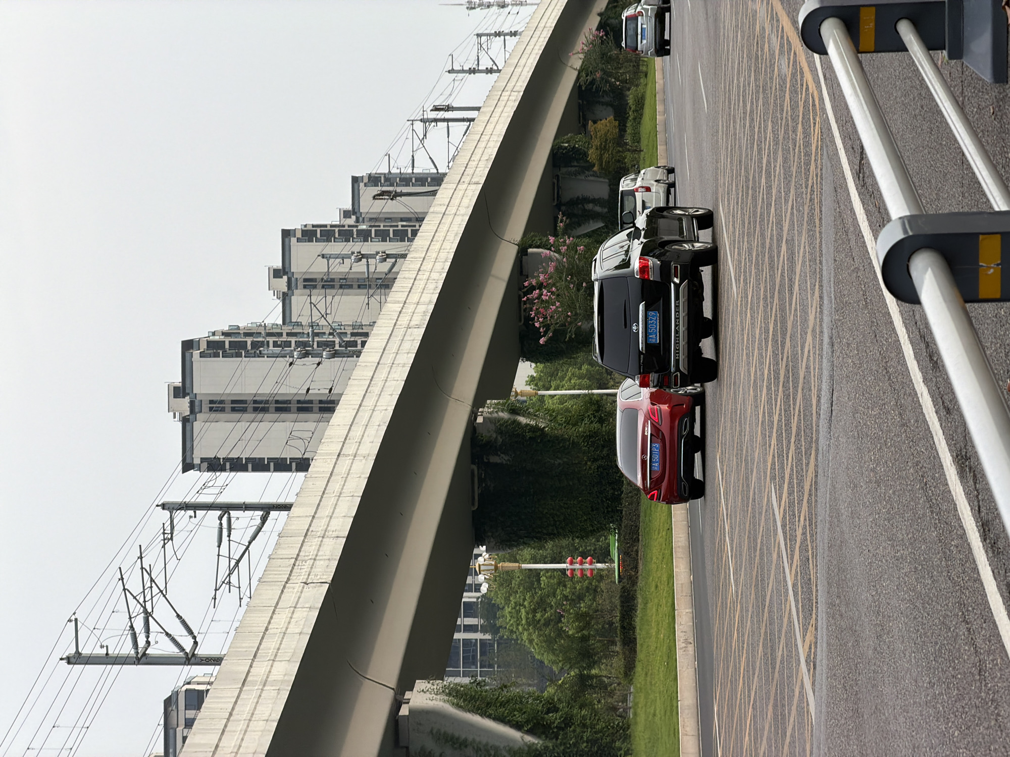 In the city of Jiangjin, China, an overpass for an elevated railway runs horizontally across the frame. The overpass is supported by concrete pillars, some of which are partially covered in green ivy. Beneath the overpass, on the right side of the image, a black SUV is driving on a multi-lane road. To its left, a red sedan is in the same lane. Further ahead, on the right, a silver SUV is also traveling on the road. The road has clear lane markings, including yellow grid lines indicating a waiting area or intersection. In the background, a row of modern, multi-story buildings with dark-tiled roofs and numerous windows stretches across the horizon. Overhead, a complex network of electrical wires and poles for the railway system is visible against a pale, overcast sky. The scene appears to be a typical urban environment with infrastructure for transportation and residential buildings.