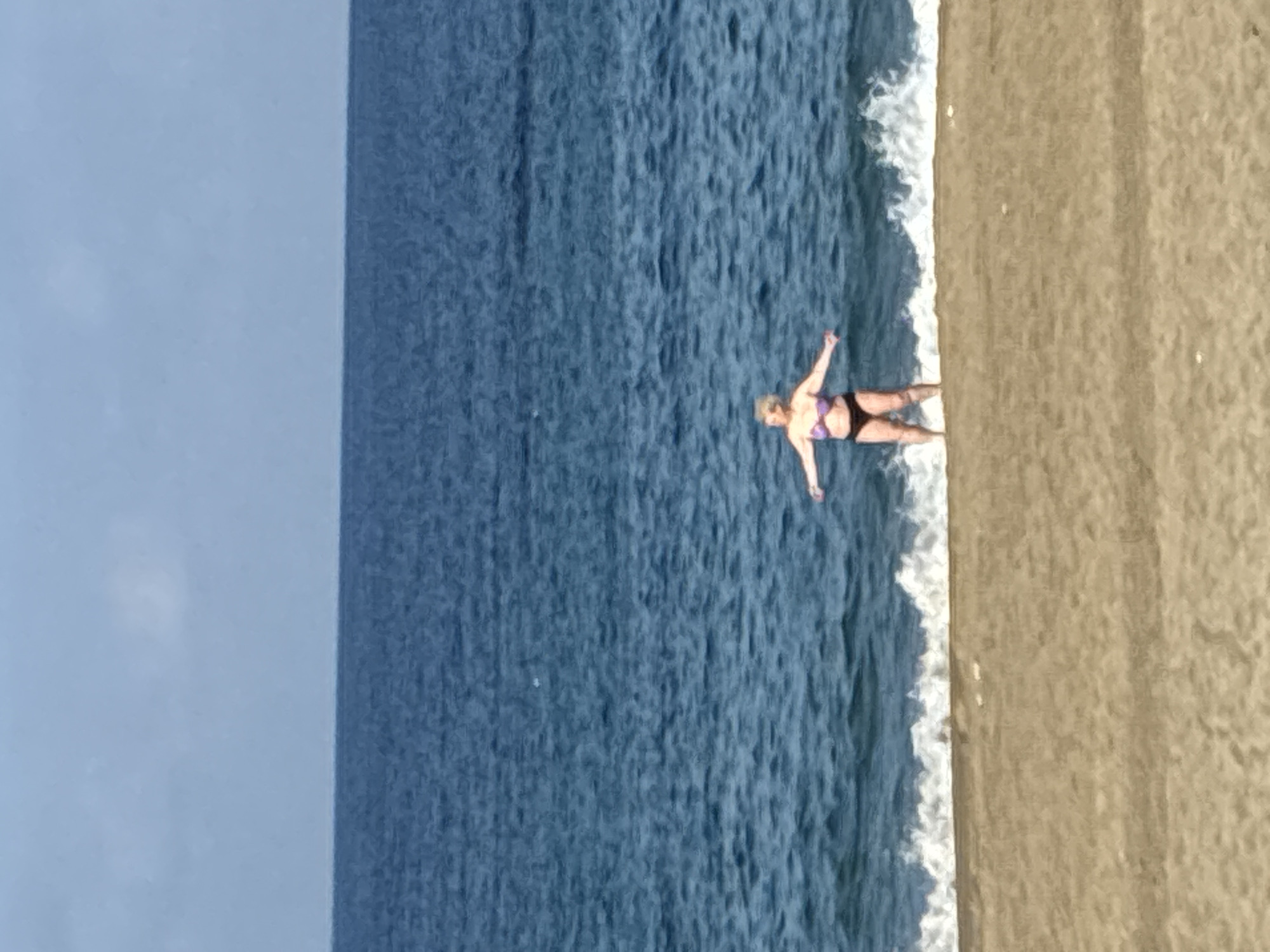 A blonde-haired woman stands in the shallow water at the edge of a sandy beach, facing the camera with her arms outstretched. She is wearing a purple bikini top and black bikini bottoms. The ocean behind her is a deep blue, with a line of white waves breaking on the shore. The sky above is a pale blue with a few wispy clouds. The beach is a light tan color, appearing smooth and unpopulated except for the woman. The image is taken from a distance, with the woman positioned in the lower middle of the frame. The overall scene depicts a sunny day at the beach. This location is likely Cleopatra Beach in Alanya, Turkey, known for its golden sands and clear blue waters.