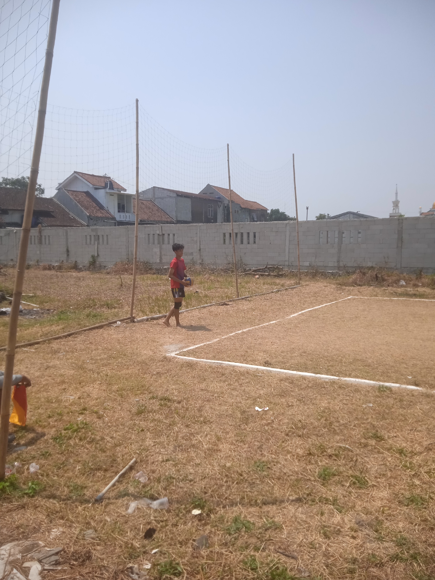 A young male in a red t-shirt and black shorts stands on a dry, grass-covered volleyball court, holding a volleyball. He is barefoot. The court is marked with white lines. A volleyball net is suspended between two wooden poles in the foreground. In the background, there is a low concrete wall with windows, and beyond that, buildings with red-tiled roofs. A mosque tower is visible in the distance. The sky is clear and bright. The scene appears to be outdoors in a semi-urban area of Klodran, Indonesia, during the daytime.