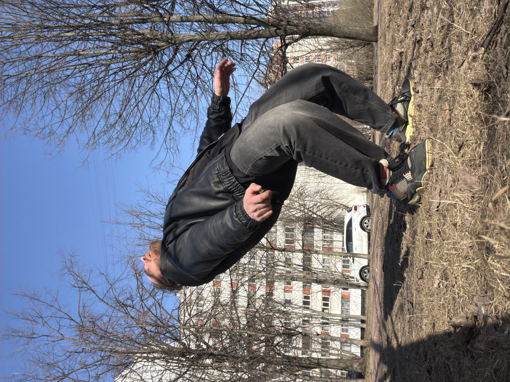 A man is depicted in an acrobatic pose outdoors. He is wearing a black leather jacket and dark grey jeans, with distinct sneakers. The background features a building with white walls and orange window frames, partially obscured by bare trees. A white car is parked in front of the building. The ground is covered in dry grass and dirt. The clear blue sky is visible through the branches of the trees. The scene is illuminated by natural daylight, suggesting a sunny day. The location is likely Saint Petersburg, Russia, based on the architectural style of the building and general environment.
