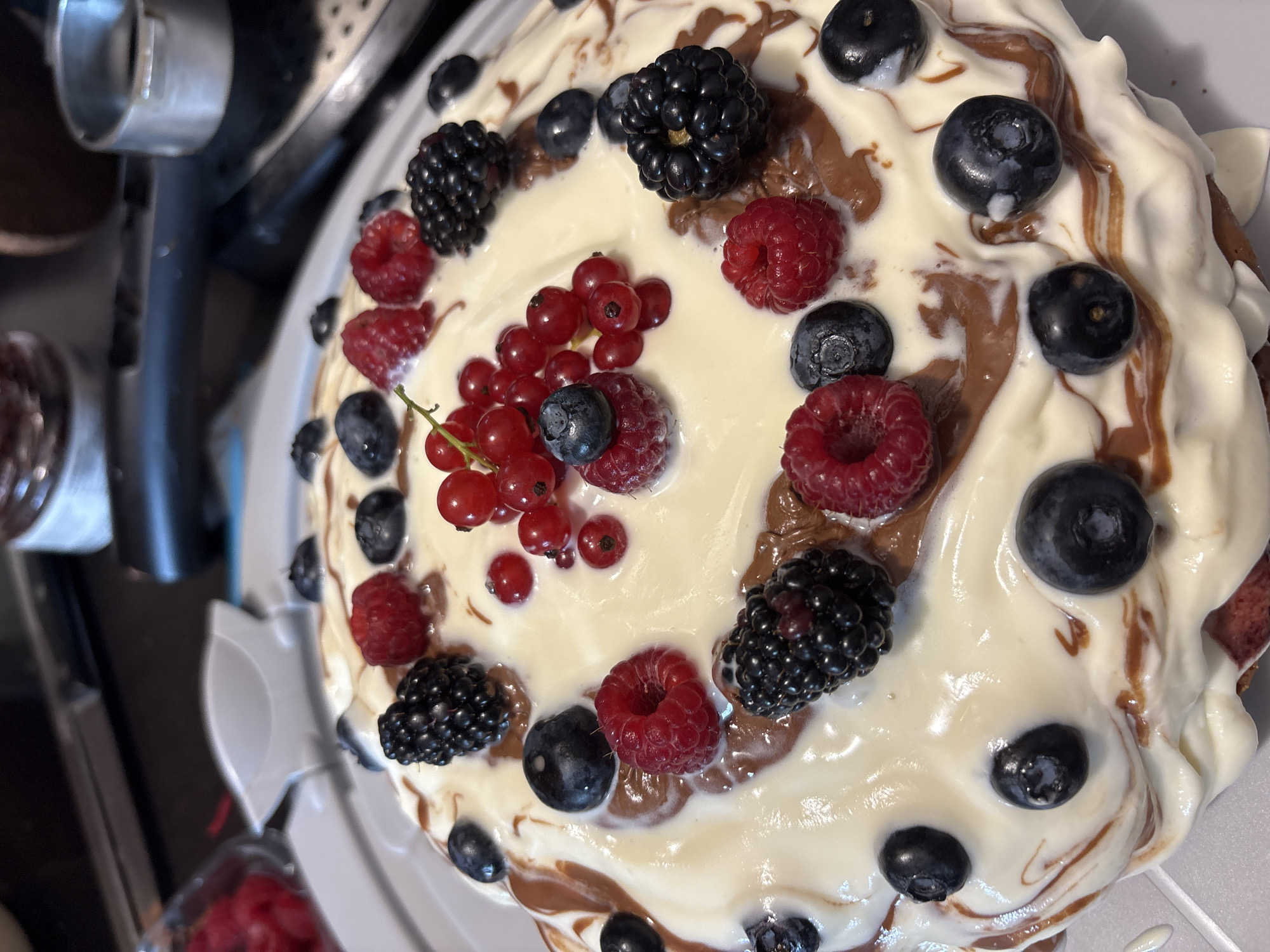 A close-up, high-angle shot captures a decadent cake topped with a swirl of white frosting, streaks of chocolate, and an abundance of fresh berries. The cake is adorned with plump raspberries, dark blackberries, bright red currants, and deep blue blueberries. The berries are scattered across the cake's surface, with a cluster of red currants forming a focal point in the center. The frosting is generously applied, creating a slightly uneven, artistic texture. The cake sits on a light gray platter, which is itself on a darker surface. In the background, out of focus, are kitchen appliances and a jar of raspberries. The lighting is bright and natural, highlighting the vibrant colors of the fruit and the creamy texture of the frosting.