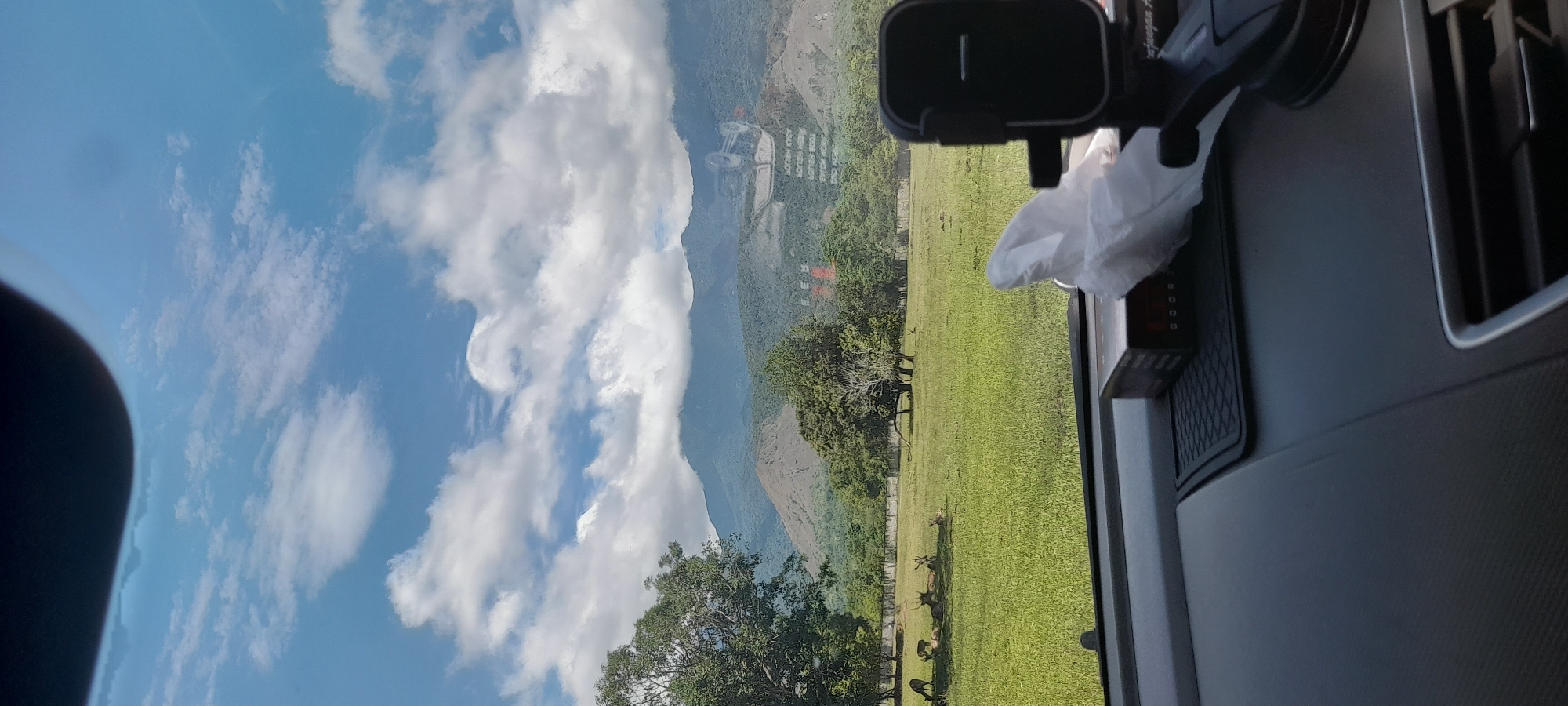 A landscape featuring a blue sky dotted with puffy white clouds and a green meadow dotted with trees is visible from inside a vehicle. A mountain rises in the background. Several deer are grazing in the meadow. A black phone mount and a small white tissue are placed on the vehicle's dashboard. The location is Unknown, Indonesia.