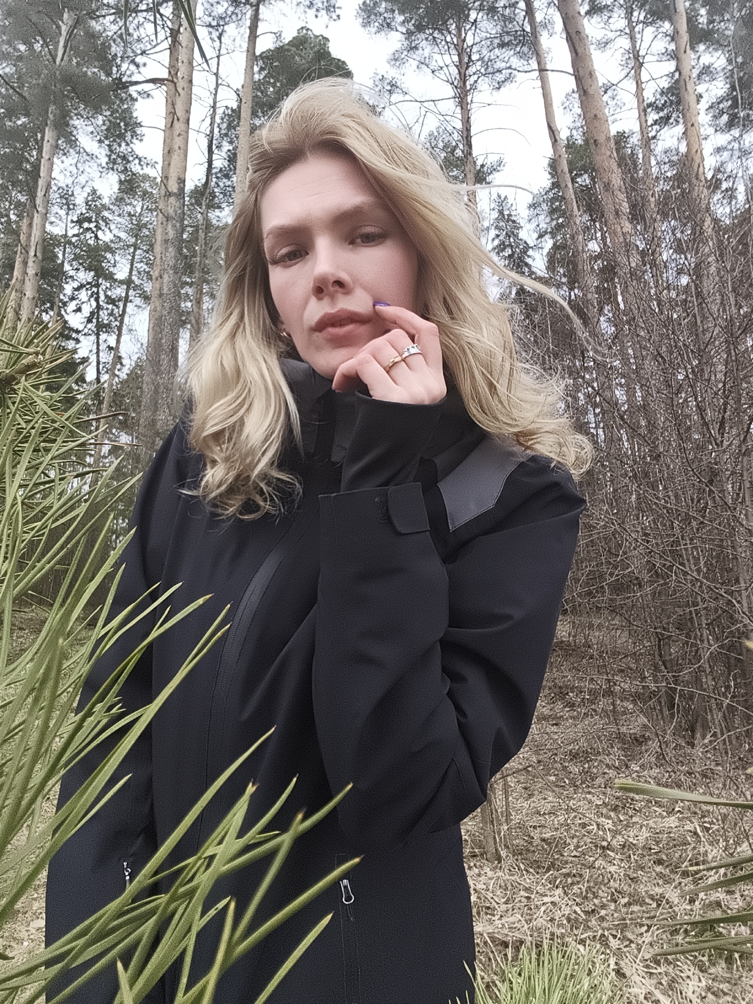A woman stands in a forest, the bare trees indicating it's likely late autumn or early spring. She is fair-skinned with wavy, blonde hair. She is wearing a black jacket, and her left hand is raised to her face, holding her chin. She has rings on her fingers, and her fingernails are painted a dark color. In the foreground, spiky green vegetation partially obscures the lower left portion of the frame. The ground is covered with dry leaves and fallen branches. The background shows a mix of bare trees and some with green foliage, under an overcast sky. The location, according to the prompt, is Izhevsk, Russia.