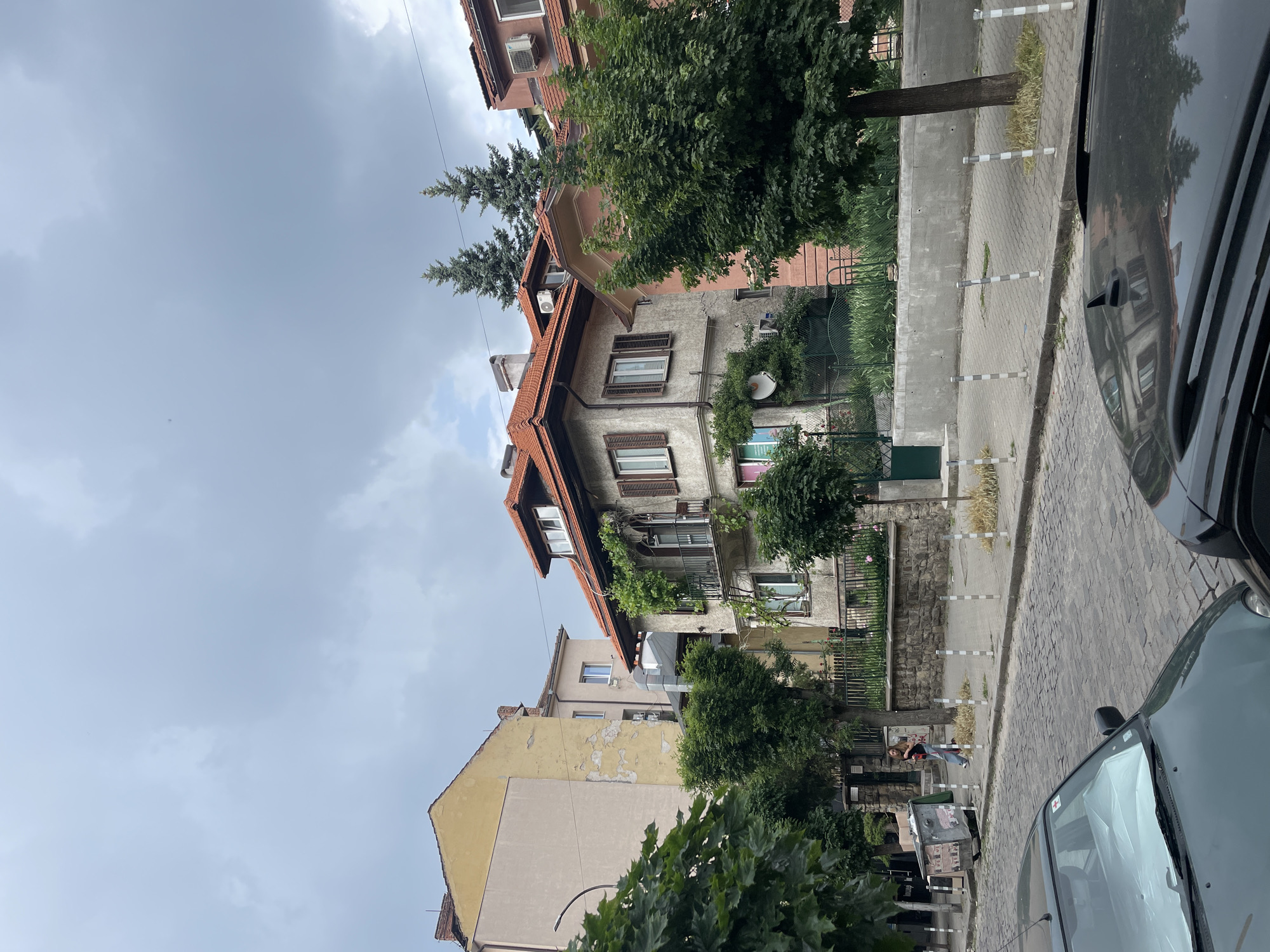 The image is a street-level shot in Sofia, Bulgaria, depicting a street scene with buildings and vehicles.
**Environment**: The sky is overcast. The street is paved with stone blocks. The architecture features older buildings with red-tiled roofs and stone or stucco facades.
**Objects**: Two cars are visible; one is green, and the other, at the right edge, is black. A trash container sits on the sidewalk. There are several trees and shrubs.
**People**: There are two people visible walking on the sidewalk.
**Subjects**: The main subject is the building with a red-tiled roof and weathered facade.