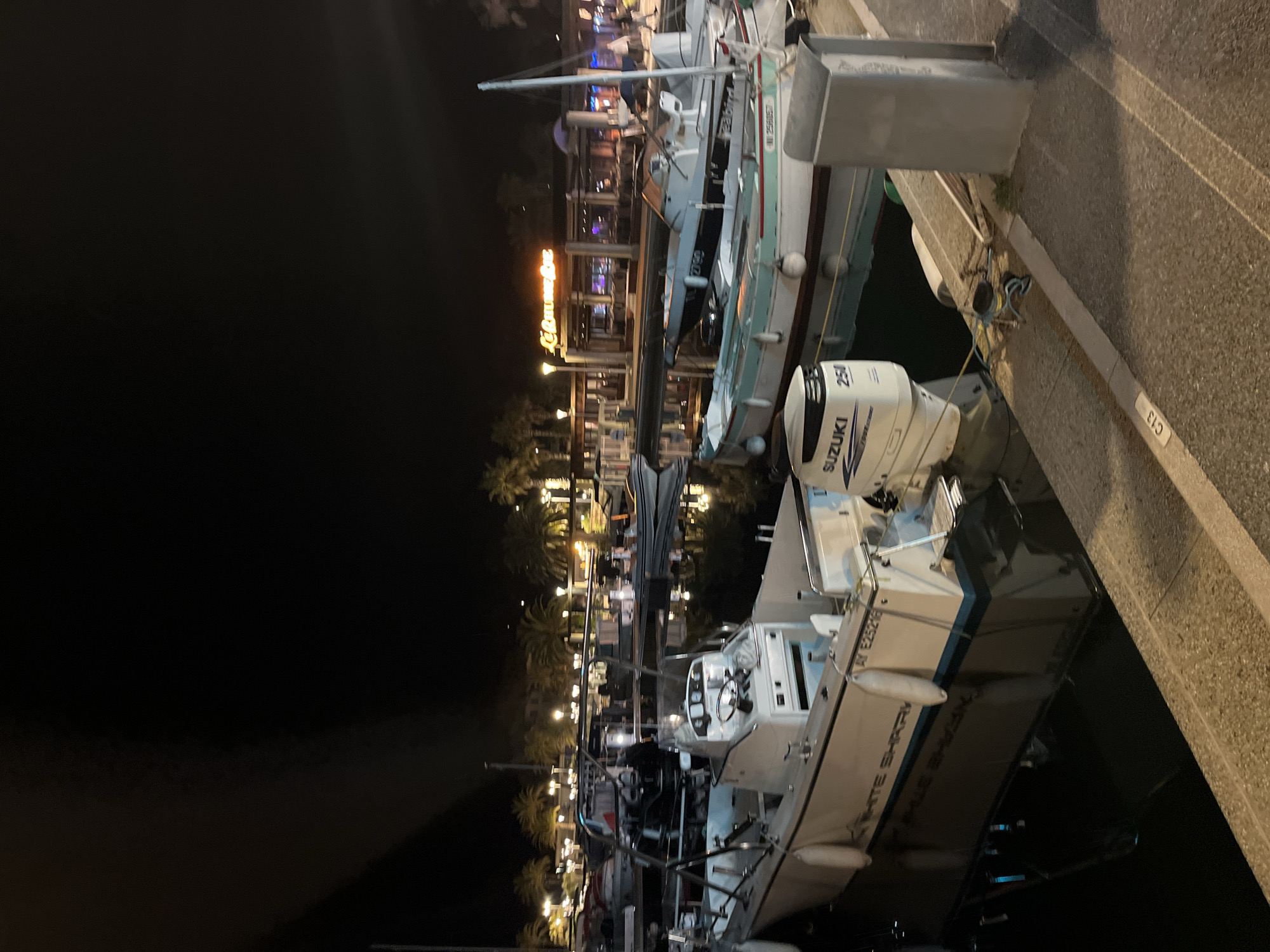 At night in Cavalaire-sur-Mer, France, boats are docked in a marina. The closest boat, partially visible, is white with the name "White Shark" inscribed. Next to it is another boat with a "Suzuki" motor, labeled "250". Beyond these, another boat is visible near a waterfront building, lit with the sign "Le Gourmet Bar". The marina and surrounding areas are lit, but the sky is dark. A concrete pier runs along the right side of the frame.