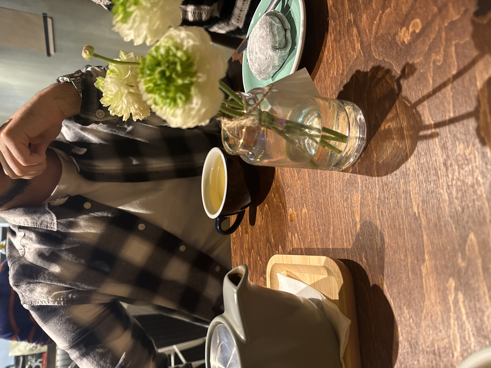 A medium-shot, angled, eye-level view depicts a wooden table at a cafe, featuring a tea set including a black cup of tea, a grey teapot on a small wooden tray, and a small glass vase containing white ranunculus flowers. A plate with a dark dessert sits nearby. A person wearing a plaid shirt is partially visible in the lower left corner, suggesting they are seated at the table. The location is Yerevan, Armenia, inferred from the setting's style and potential tea-serving traditions. The scene is illuminated by natural light, creating distinct shadows on the wooden table.