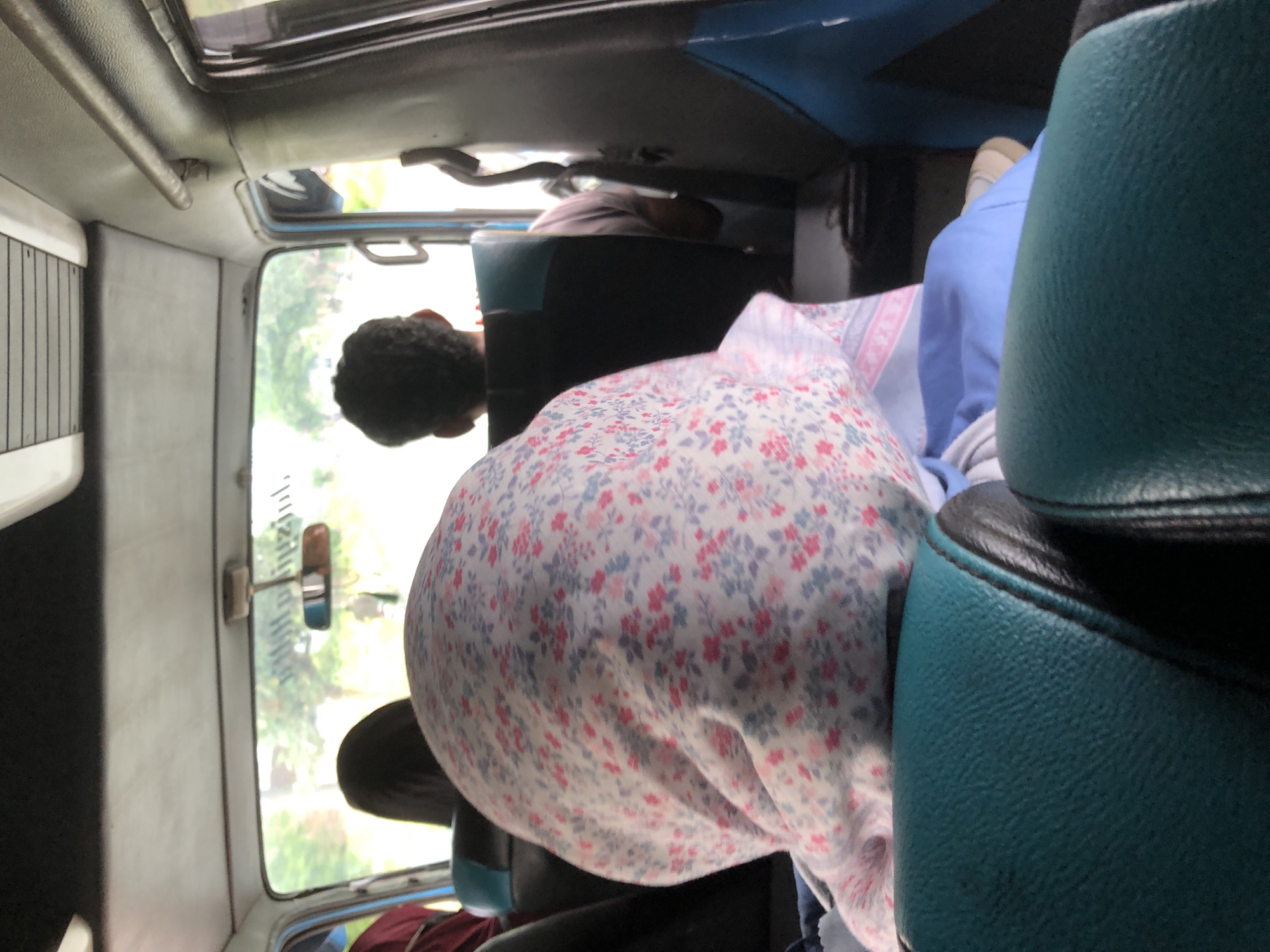 The image, taken in what appears to be a moving vehicle, perhaps a small bus or van, shows the interior of the vehicle with passengers. The scene is dominated by the back of a person's head, covered by a hijab-like garment with a small floral pattern, possibly a pink and blue design. To the right of this person is a blue seat, likely vinyl or leather. The vehicle's windows offer a blurred view of the outside, potentially showing trees or vegetation. There is also another person further up in the bus but its hard to see details. The image was captured in Putoh Sa, Indonesia.