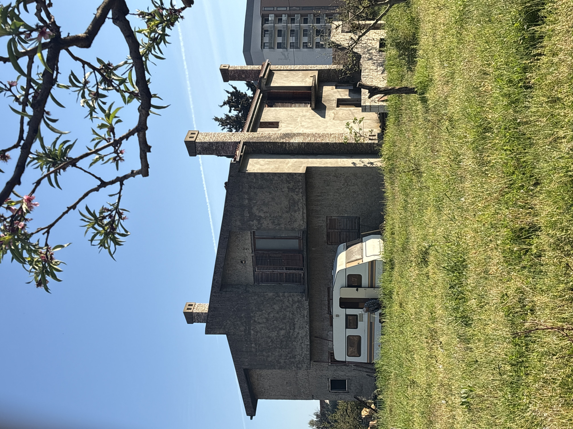 An eye-level, outdoor shot shows a two-story, gray concrete house with brick trim in the city of Bar, Montenegro. The house has three brick chimneys and two windows on each story with brown louvered shutters. To the right of the house is a camper parked on a green lawn and a person, presumably the camper owner, standing next to the camper. In the background is a white multi-story apartment building. To the left is a tree branch with small pink blossoms set against a blue sky with white vapor trails. The ground appears to slope slightly upward to the right. The image is rotated, so the house and camper appear to be on their side.