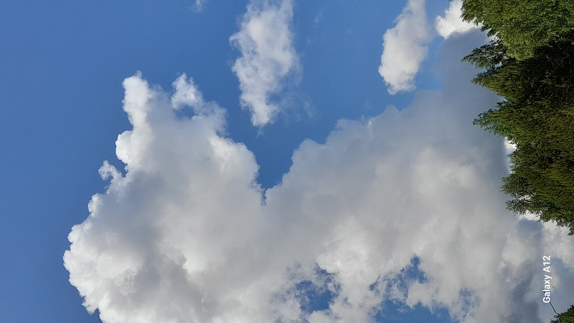 The image shows a view of the sky with white clouds and a bright blue background. The lower right corner has a blurred green area which appears to be tree foliage. The words "Galaxy A12" are also visible in the lower right corner, suggesting the image was taken with that phone model. The specific location within the city of Марковский, Russia cannot be determined from the visual information provided. There are no visible people, actions, or interactions present in the image.