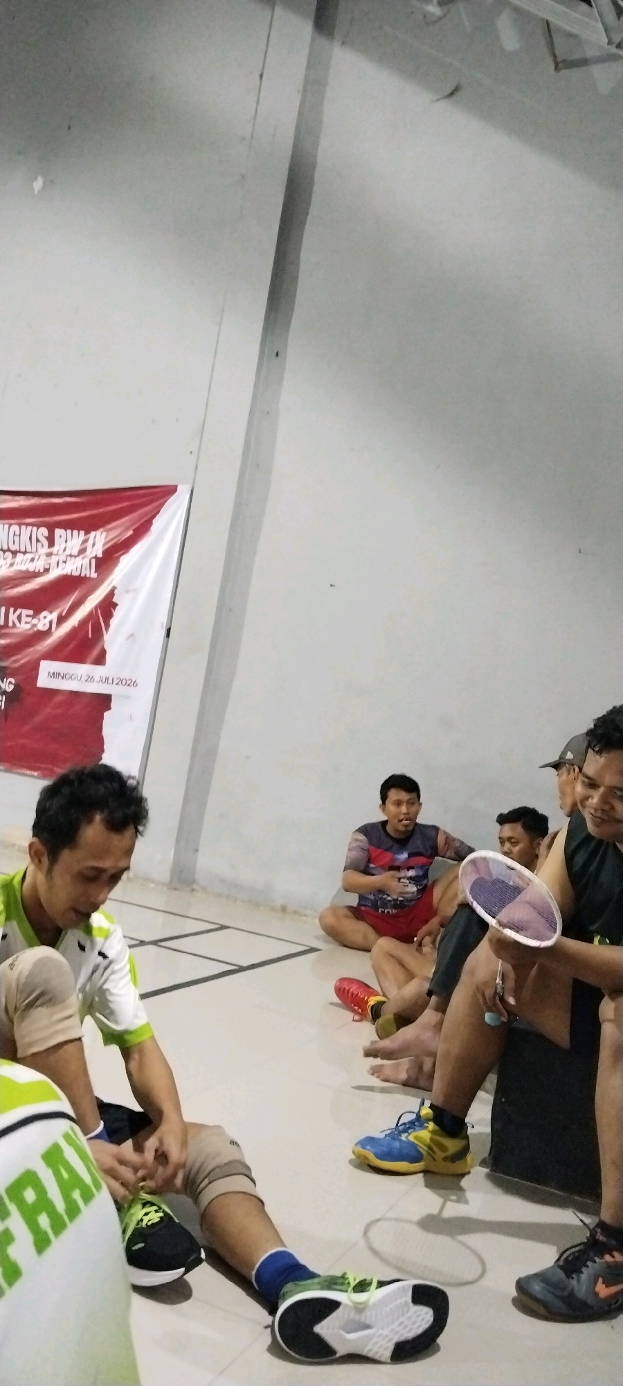 The image depicts a group of young adult males sitting on the floor of what appears to be an indoor badminton court, likely in Kliris, Indonesia. The court floor is marked with white lines. A large banner is visible on the left side, with text indicating an event or organization, including "RW IX" and "Minggu, 26 Juli 2026".
Four individuals are prominently visible. In the foreground left, a man in a green and white shirt and knee support is looking down, with his hands resting on his knees. Next to him, to the right and slightly further back, another man is sitting with a badminton racket in his hand. He is wearing a dark shirt with a colorful design and red shorts. Behind him, two other men are seated. One is wearing a dark t-shirt and red shorts, and the other is wearing a dark shirt and a cap. The lighting is somewhat dim, suggesting an indoor setting. The overall atmosphere appears relaxed, with the men taking a break.
