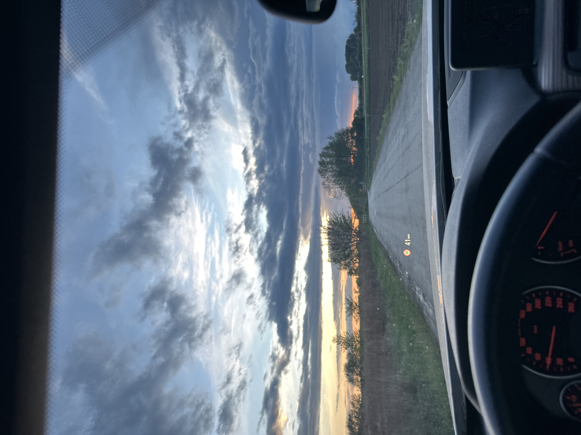 The image shows a view from inside a car driving on a rural road. The sky is filled with clouds and the setting sun is visible in the horizon. The road is lined with fields and trees. A speed limit sign is visible on the side of the road. The car's dashboard and steering wheel are visible in the foreground.