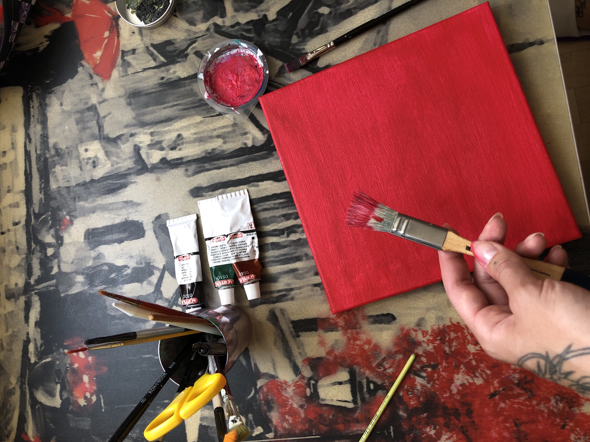 An overhead, eye-level shot shows an artist's workspace, with a red canvas occupying the lower right quadrant. A hand is holding a paintbrush with red bristles to the canvas. To the left of the canvas, tubes of acrylic paint are visible, along with a cup filled with bright red paint and a small container of black material. A yellow pair of scissors and a collection of paintbrushes are in a metal cup in the upper left. The background appears to be an abstract painting with muted tones of gray, black, and red. The scene does not provide any specific details about the city of Boryspil, Ukraine, or any landmarks that would indicate its location.