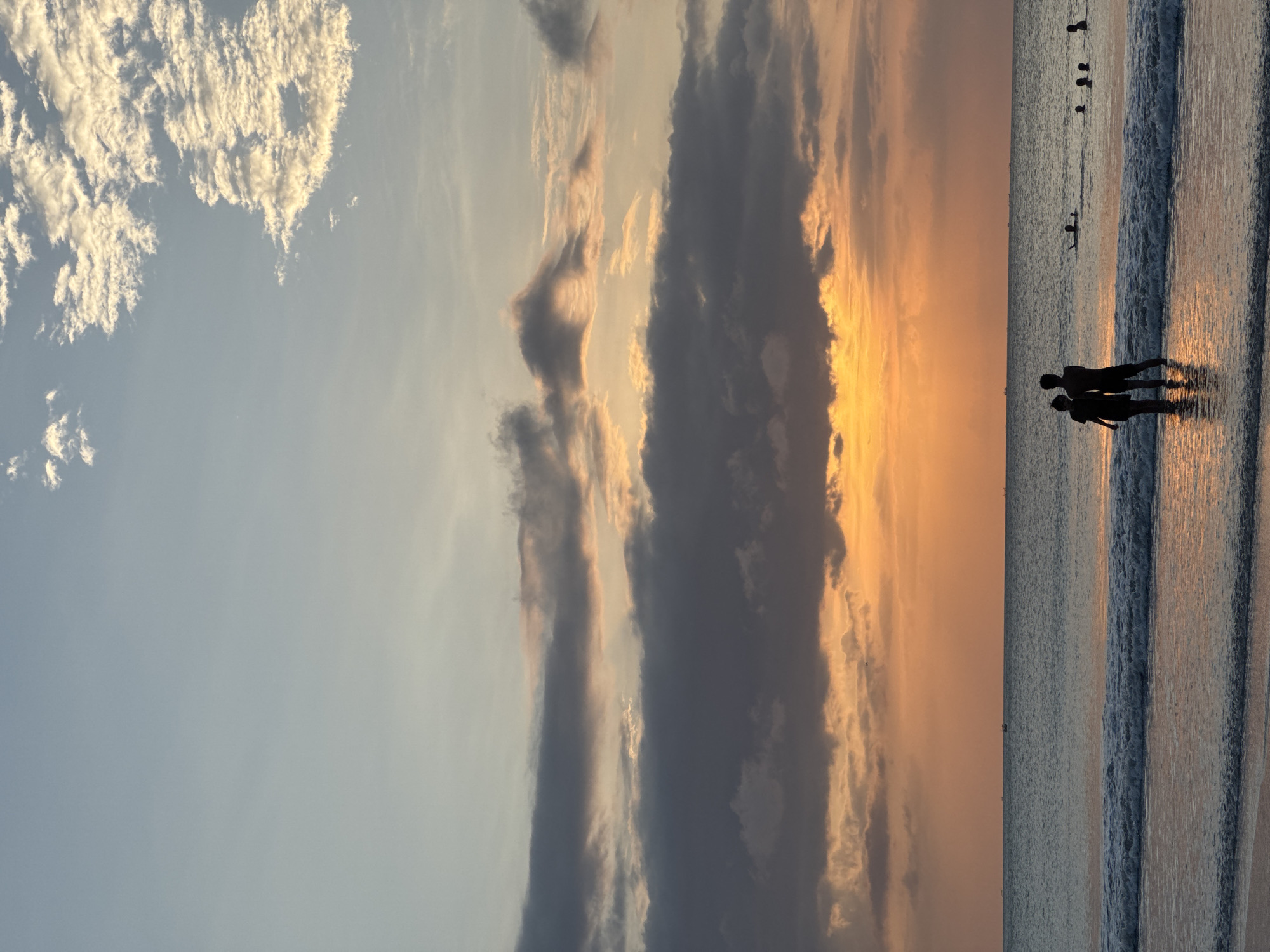 A vertical, low-angle shot captures a serene beach scene at sunset in Kuta Selatan, Indonesia. The sky is a gradient of soft blues, transitioning to warm oranges and yellows as the sun dips below the horizon, its light diffused by scattered clouds. The ocean is calm, with gentle waves lapping at the shore.
In the foreground, a silhouetted couple stands close together in the shallow water, facing the sunset. Their figures are dark against the illuminated water. Further out in the ocean, several other individuals are visible, some swimming and others wading. A thin line of distant boats is visible on the horizon. The wet sand in the foreground reflects the warm hues of the sky. The overall impression is one of peacefulness and natural beauty.