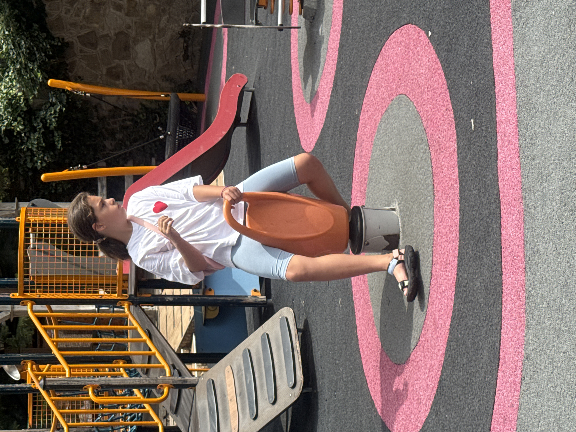 A young girl with brown hair in a ponytail is playing on a playground. She is wearing a white t-shirt with a red heart on it, light blue shorts, and black and white sandals. She is sitting on an orange, plastic seat attached to a spinning metal pole, with her legs outstretched. Her left hand is holding onto the orange seat, and her right arm is bent as she looks to her left. The playground has a black rubberized surface with pink and gray circular designs. In the background, there is a yellow slide, a section of a red slide, and a yellow metal structure with stairs. The scene appears to be outdoors during the daytime, with sunlight casting shadows. The girl's facial expression is not clearly discernible, but she appears to be engaged in play. The location is identified as Yalta, Ukraine.