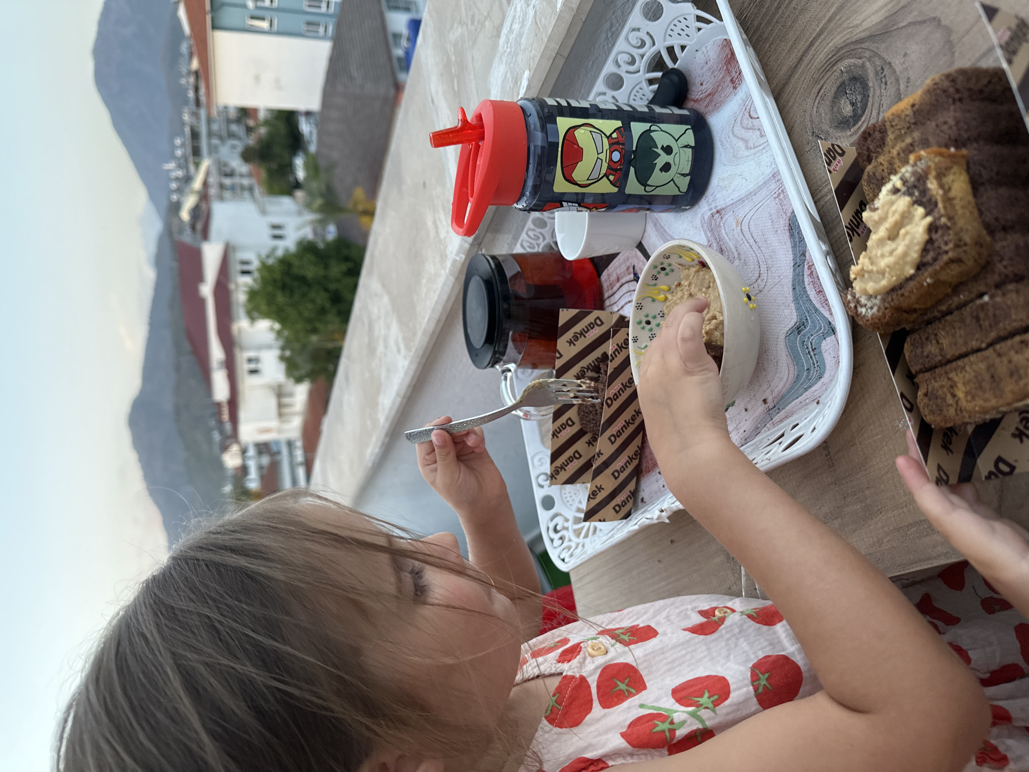A child with brown hair, wearing a light-colored dress with a tomato pattern, sits at a table on an outdoor terrace and eats from a floral-patterned bowl using a fork. On the white plastic serving tray are a jar of tea, a child's water bottle with superhero characters, and cut slices of Dankek brand cake. The table is on a balcony overlooking a cityscape with white buildings and a distant mountain range. The scene is likely set in Fethiye, Turkey, given the product label "Dankek," which is popular there.