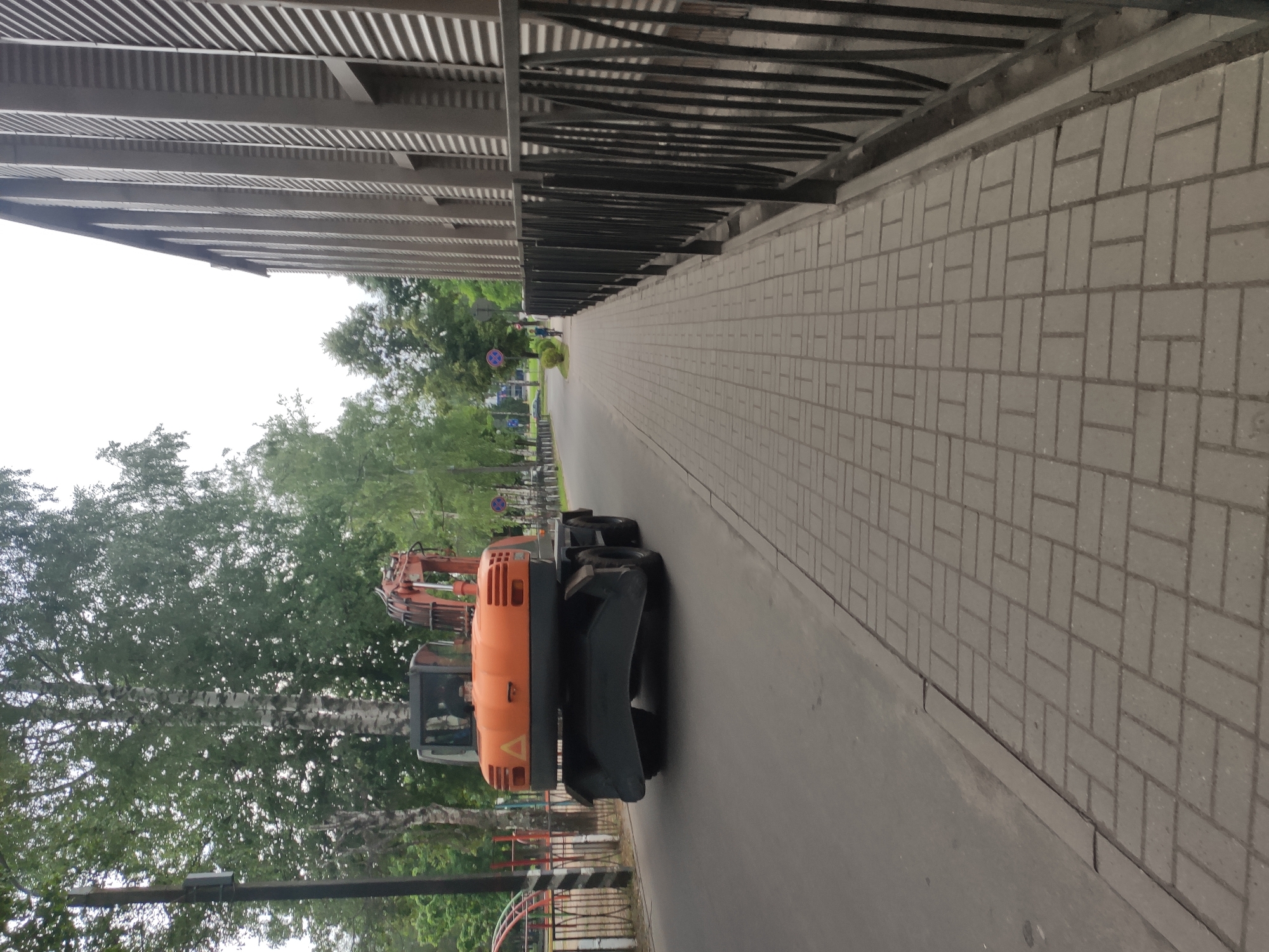 An orange excavator is driving on a paved road in Navapolatsk, Belarus. The excavator is in the left lane of the road and is moving away from the viewer. To the right of the road is a sidewalk paved with rectangular bricks. A black metal fence runs along the edge of the sidewalk. In the background, there are trees and a building with a corrugated metal facade. The sky is overcast. A person on a bicycle is visible on the sidewalk in the distance.