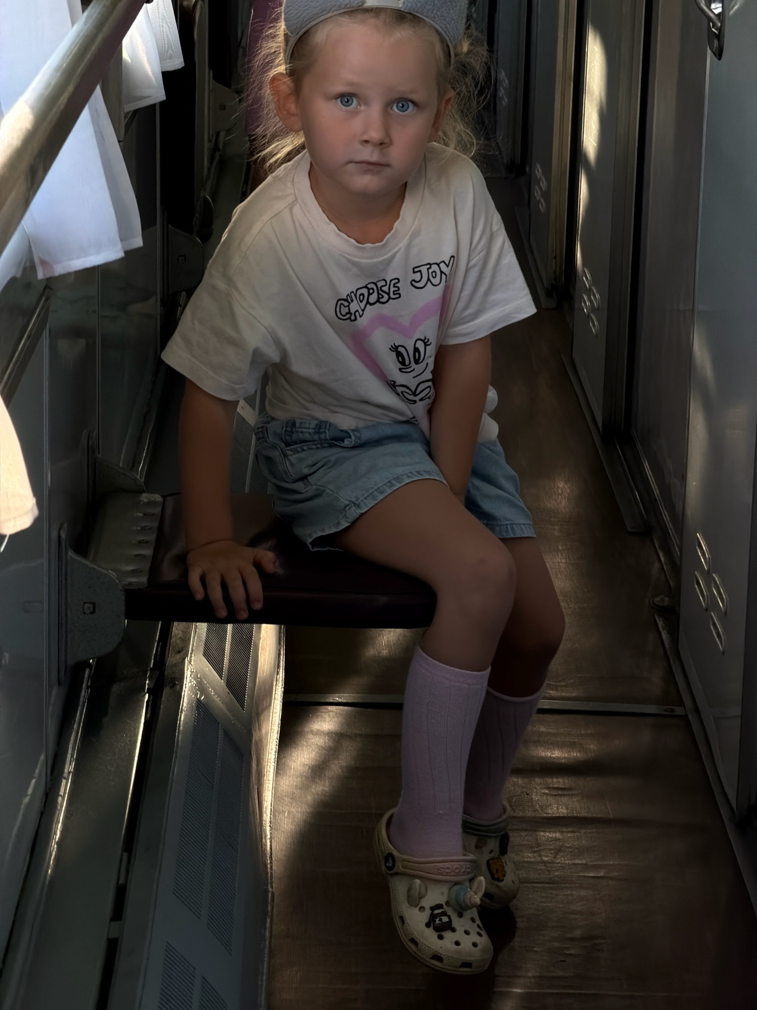 A young girl with blonde hair and blue eyes, wearing a grey headband, a white t-shirt with "CHOOSE JOY" and a heart graphic, denim shorts, light purple socks, and white Crocs with charms, sits on a dark red bench inside what appears to be a train compartment in Il'men', Russia. Sunlight streams in from the right, casting shadows on the wooden floor and the side of the compartment, suggesting it is daytime. The girl looks directly at the camera with a neutral expression. The setting includes a metal railing on the left, white fabric hanging, and the interior of a train carriage with grey walls and doors. The girl's hands rest on the bench.