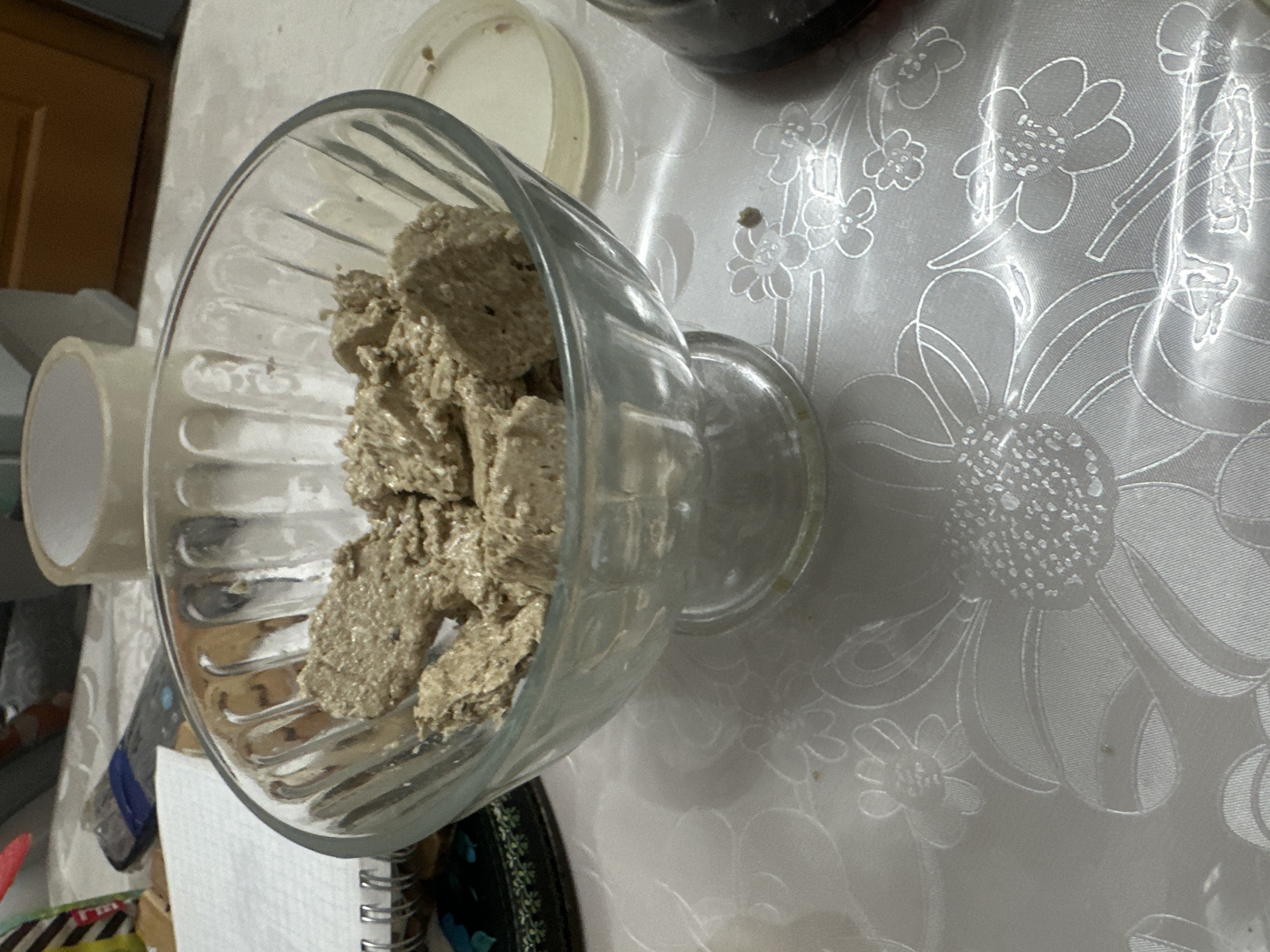A glass dessert bowl filled with halva sits on a white tablecloth with a floral pattern. The halva is broken into irregular chunks and appears to be a light brown color with small dark specks. To the right of the bowl is a white plastic lid, and further to the right is a dark, cylindrical object, possibly a jar. In the background, a roll of tape and a remote control are visible on a surface that appears to be a table or desk. The overall scene suggests a casual setting, likely in a home or dining area. There is no indication of a specific location within Загородный, Russia, nor are there any people present.