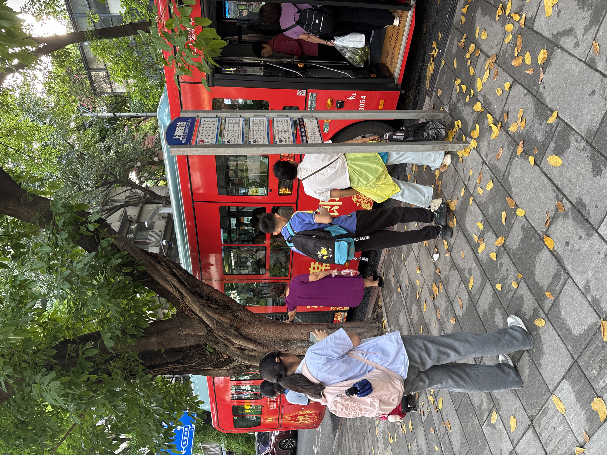 The image shows a bus stop on 龙黄公路 (Long Huang Highway) in the Nan'an District of China. A red double-decker bus is stopped, with passengers boarding. Several people are standing in line to board the bus. The bus stop sign reads "上新街站" (Shang Xin Jie Station) and "BUS STOP SHANG XIN JIE". Leaves are scattered on the pavement. A large tree is next to the bus stop.