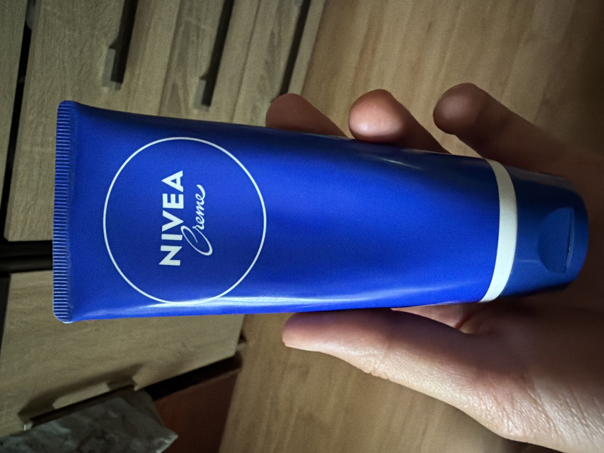 A close-up shot shows a person's hand holding a tube of Nivea Creme. The tube is bright blue with the Nivea logo in white, featuring the word "NIVEA" in bold, capitalized letters above "Creme" in a script font, all enclosed in a white circle. The tube has a ribbed cap at the bottom and a white band just above it. The hand is positioned on the right side of the frame, supporting the tube. The background is blurred and appears to be an indoor setting with wooden flooring and what looks like a dresser or cabinet with drawers. The lighting suggests an indoor environment with some shadows. The overall scene is static and focused on the product being held.