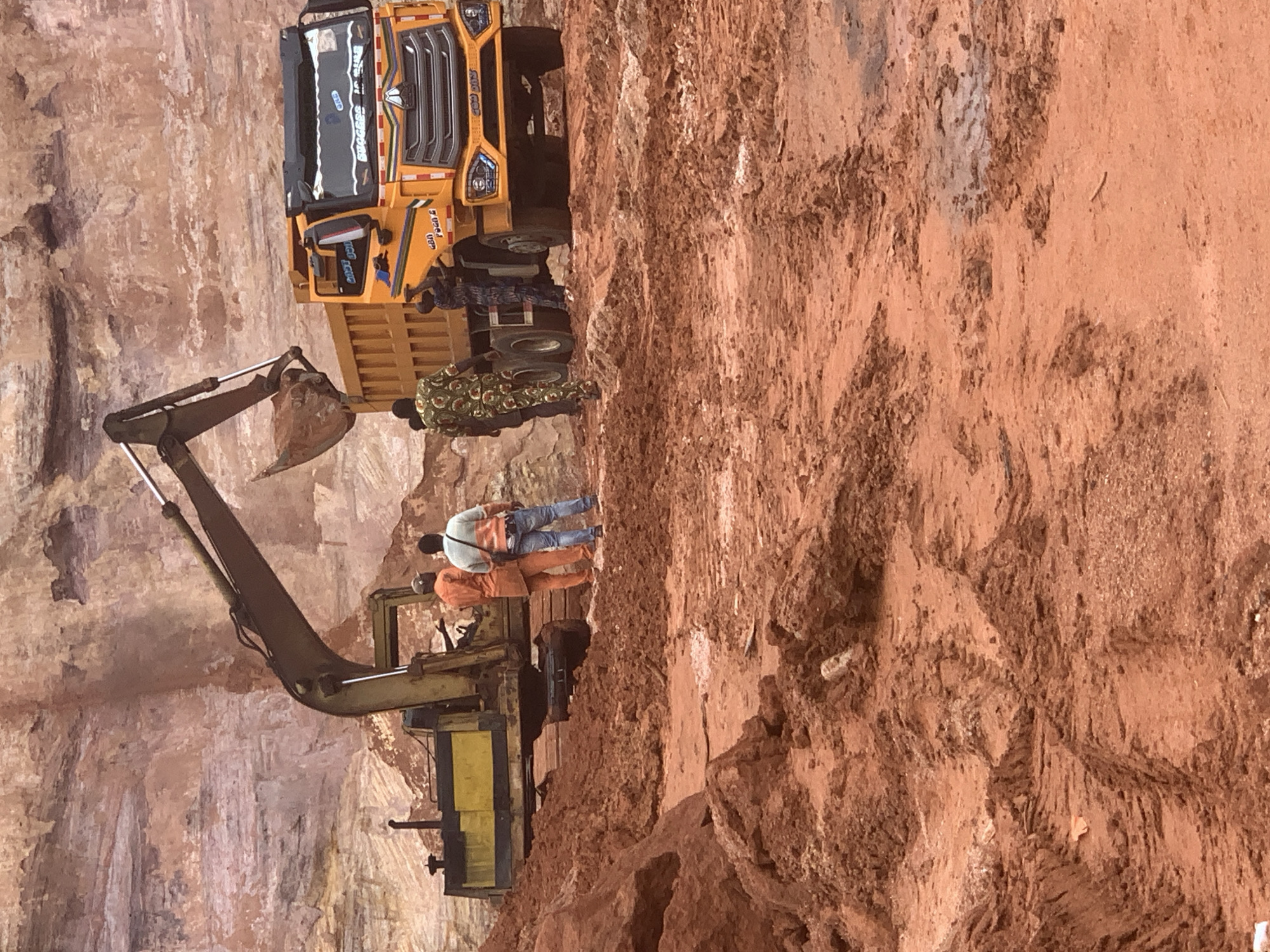 The image depicts an open-pit mine in Ijesha-Ijebu, Nigeria. The foreground is dominated by reddish-brown soil with visible tire tracks from heavy machinery. In the midground, a large yellow excavator is positioned, with its arm extended over a bright yellow dump truck. Several individuals are present. Two men in the center appear to be wearing orange and light-colored shirts respectively. Further to the right, another individual is seen near the dump truck. The background shows a steep, layered rock face characteristic of an excavation site. The overall scene suggests ongoing mining operations.