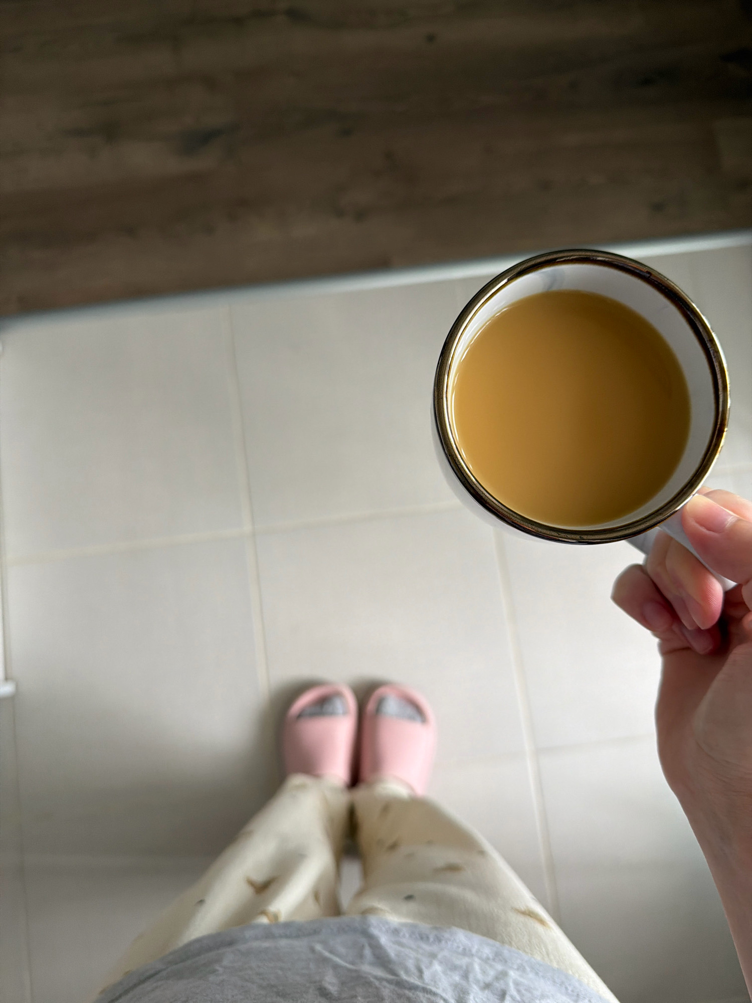 A person's hand holds a white mug with a gold rim filled with a light brown liquid, likely coffee, in an indoor setting with light-colored tiled flooring. The person is wearing pink slippers and light-colored patterned pants. The background shows a wooden surface. The scene is viewed from a top-down perspective. The image does not contain enough information to determine the location in the city of Kyiv, Ukraine.