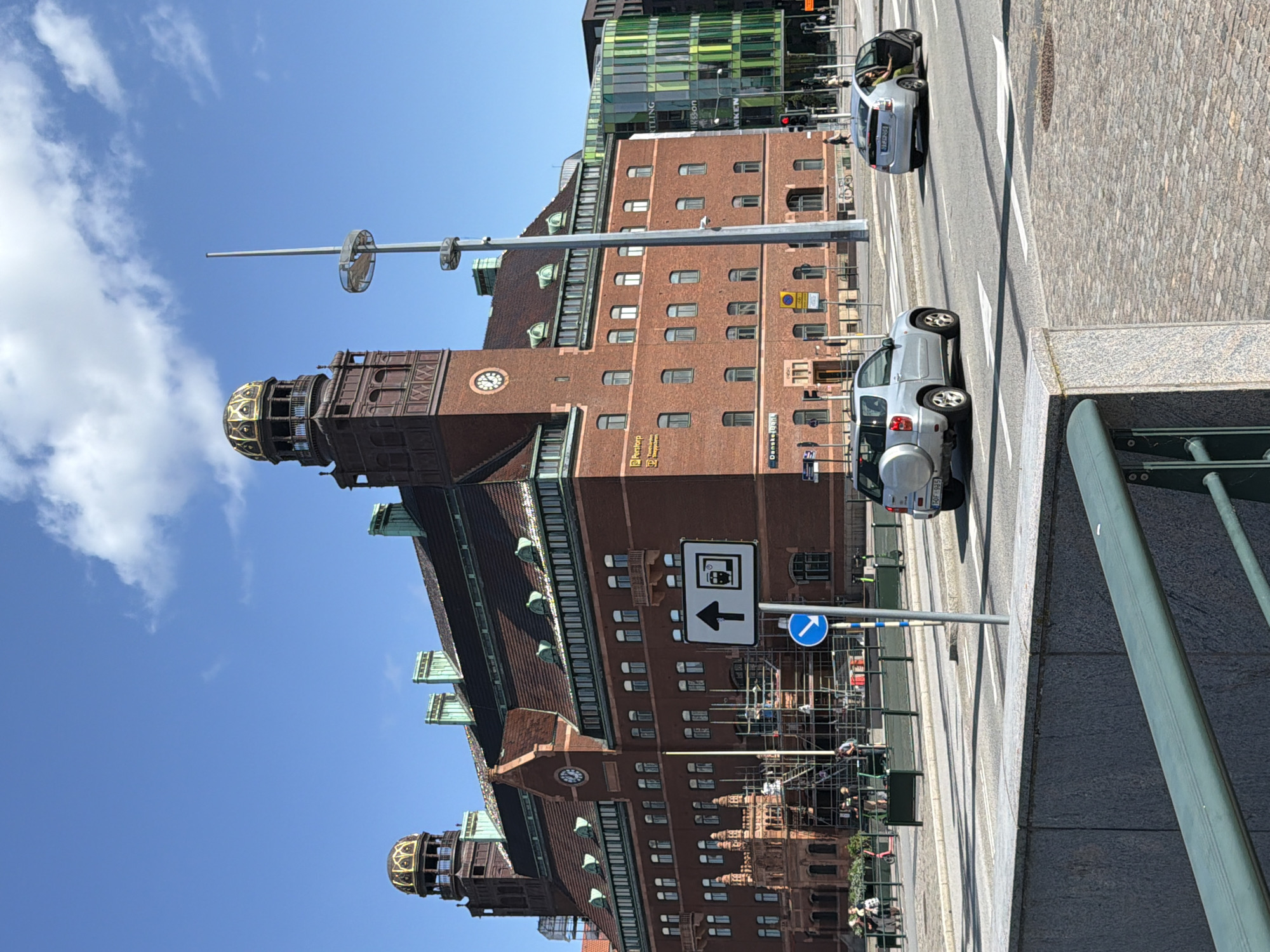 The image captures a street view in Malmö, Sweden, featuring a prominent red brick building with copper-colored domes and green accents. The building appears to be a historical structure, possibly an administrative or bank building, with multiple windows and ornate architectural details. In front of it, a street with traffic signs, including an arrow pointing forward with a symbol of a tram, is visible. Two cars are on the street; one silver SUV parked near the intersection and a silver sedan with its driver's side door open, with a person inside wearing a green outfit. In the background, a modern, multi-colored building with green and blue glass facades stands out. The sky is blue with scattered white clouds, and the lighting suggests it is a sunny day. The scene is bustling with pedestrian activity and suggests a typical urban environment.