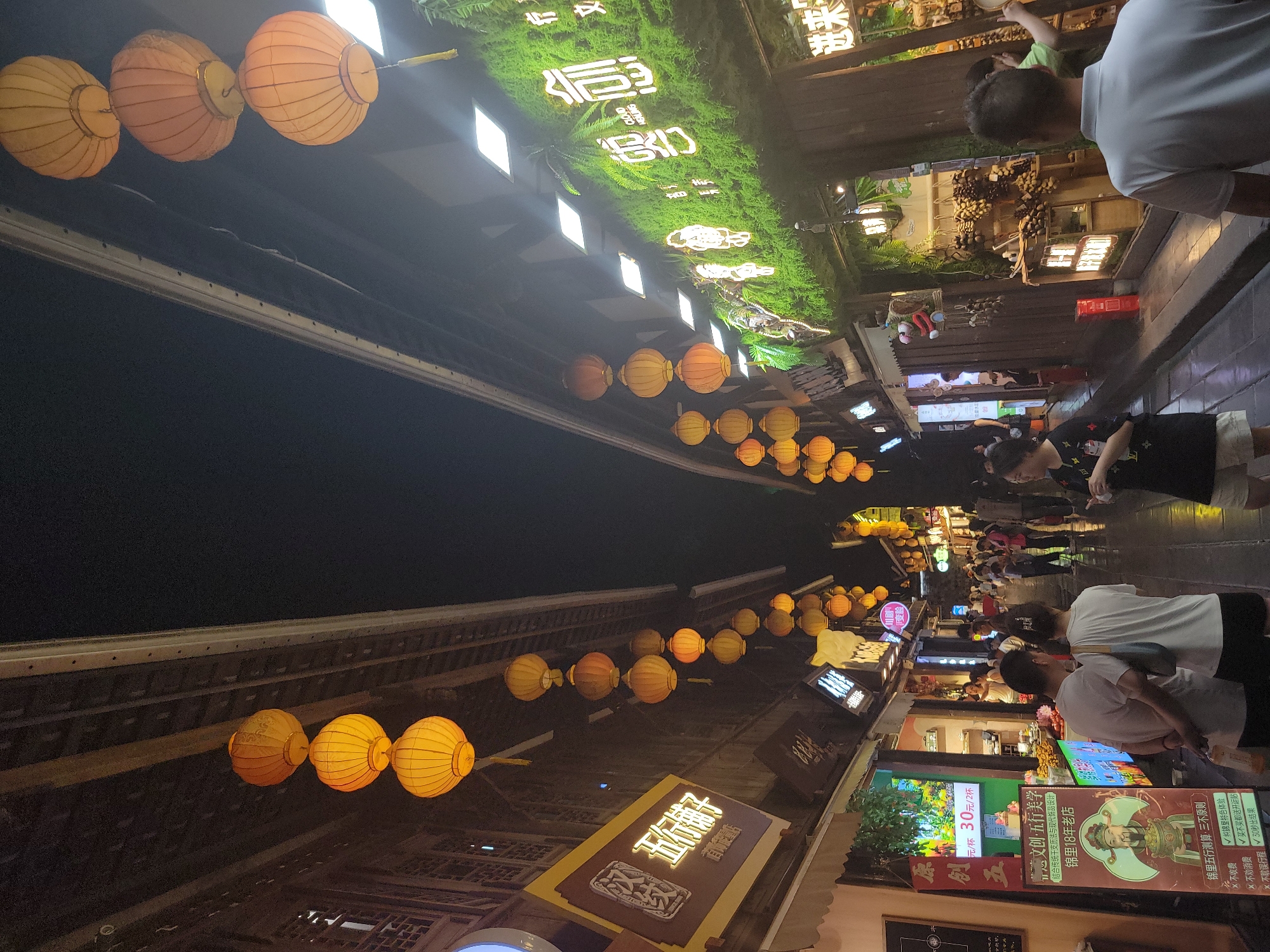 The image depicts a bustling street scene at night in the Wuhou District of China, likely a commercial or entertainment area. Traditional Chinese lanterns, primarily orange and yellow, are suspended overhead, illuminating the narrow street lined with buildings on both sides. These buildings feature wooden facades and illuminated signs, some with Chinese characters and others with modern neon lettering, including one sign with glowing green foliage and text.
Several people are visible walking along the street, some in small groups and others individually. They are dressed in casual attire. Some storefronts display goods, such as fruits or other merchandise. The ground appears to be paved, and there are reflections visible, suggesting it might be wet. The overall atmosphere is lively and vibrant, characteristic of a night market or a popular tourist destination.