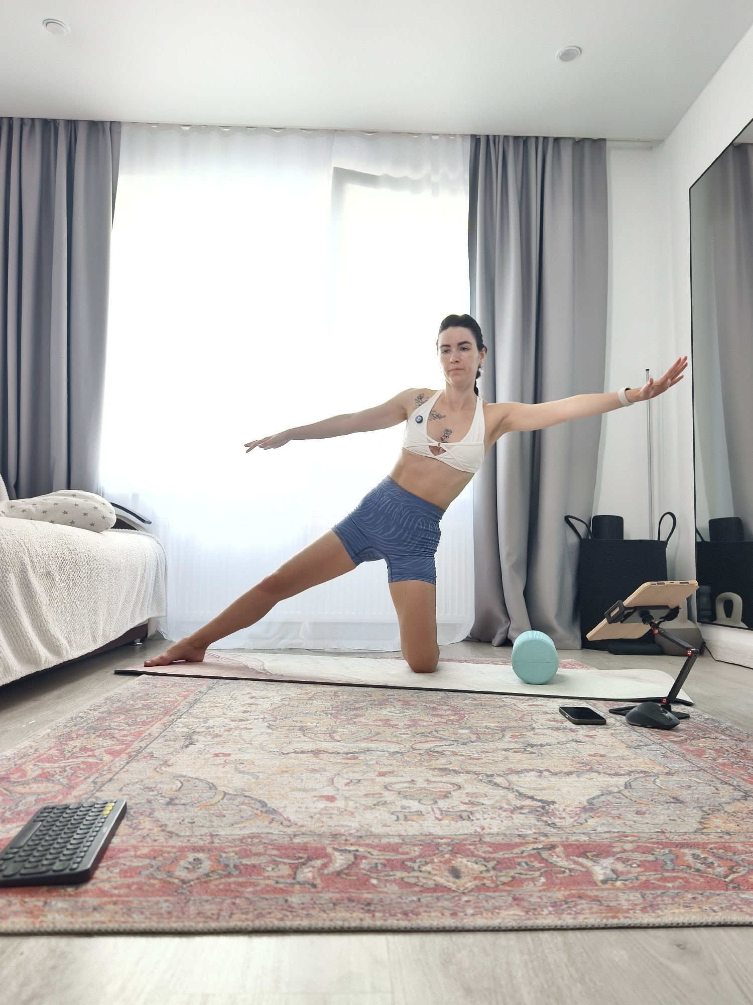 The image depicts an indoor scene in what appears to be a bedroom or a multi-purpose room, possibly in Tyumen, Russia. A woman is performing a yoga or Pilates exercise on a mat. She is kneeling on her right knee, with her left leg extended to the side, and her arms are outstretched. She is wearing athletic attire: a white sports bra and blue patterned shorts.
In the background, there is a bed with white bedding and a patterned bolster pillow. A large window is covered by gray curtains and sheer white drapes, allowing natural light to illuminate the room. To the right, a tall mirror reflects part of the room, and black storage baskets are visible.
On the floor, several objects are placed: a light blue yoga block, a smartphone, a tablet mounted on an adjustable stand with a black base, and a black wireless keyboard. The floor is covered with a large, intricately patterned rug. The overall setting suggests a domestic environment adapted for fitness activities.