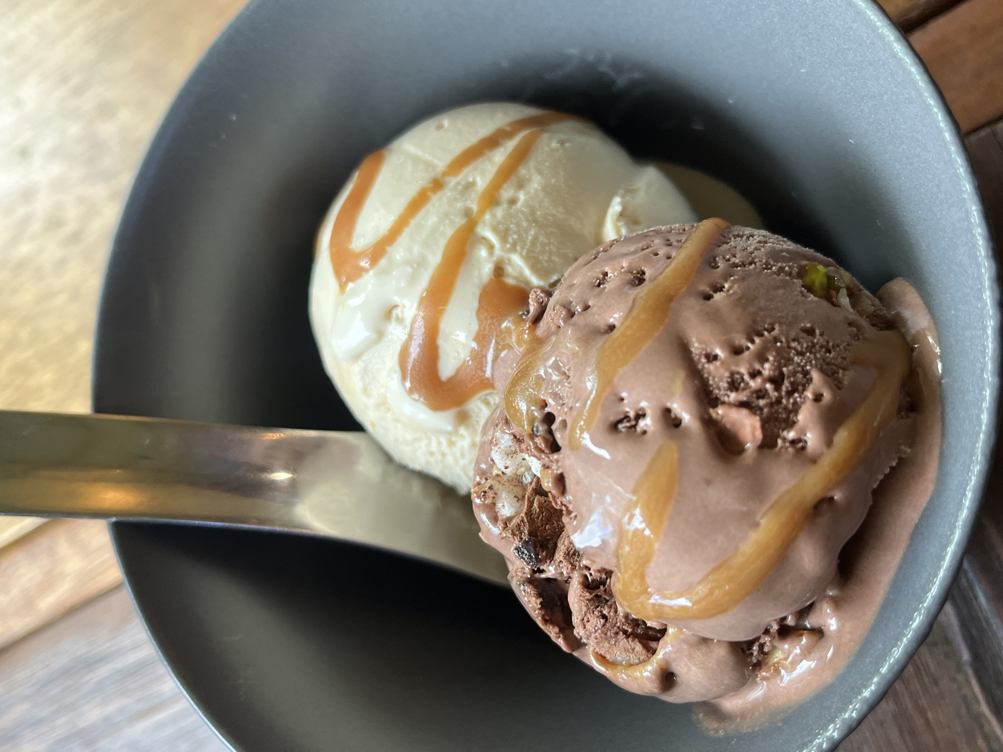 A close-up, overhead shot captures two scoops of ice cream in a dark gray bowl. The scoop in the foreground is chocolate, with streaks of caramel sauce drizzled on top. Small chunks of what appear to be chocolate chips or nuts are visible within the chocolate ice cream. Behind it, a scoop of vanilla or cream-colored ice cream is also adorned with caramel drizzle. A metallic spoon is partially visible, resting in the bowl between the two scoops. The bowl sits on a wooden surface, suggesting a restaurant or home setting. The image appears to have been taken in ตำบลเกาะพะงัน, Thailand.