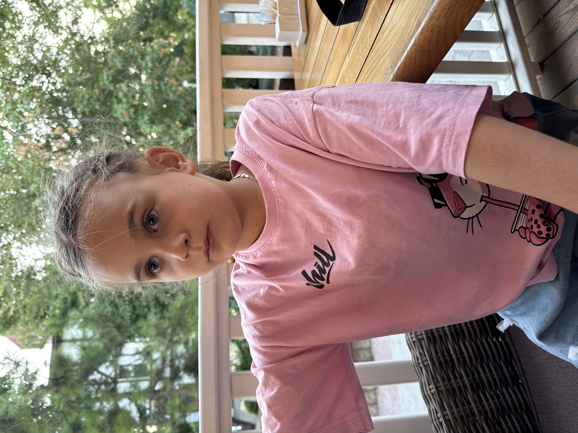The image shows a young girl from the shoulders up, sitting at a wooden table outdoors. She has long, light brown hair pulled back in a messy bun, and her gaze is directed towards the camera. She is wearing a light pink t-shirt with a black graphic design that appears to be a cartoon character and some text. She is also wearing a silver necklace.
The setting appears to be a porch or patio with a railing in the background and lush green trees and foliage visible. The lighting suggests it is daytime, possibly late afternoon or early evening, with the sun filtering through the leaves, creating a soft, natural glow. The overall mood seems relaxed and casual. The text on her shirt is partially visible and reads "chill". The girl's expression is neutral, looking directly at the viewer. The scene is well-lit and appears to be a pleasant outdoor setting.