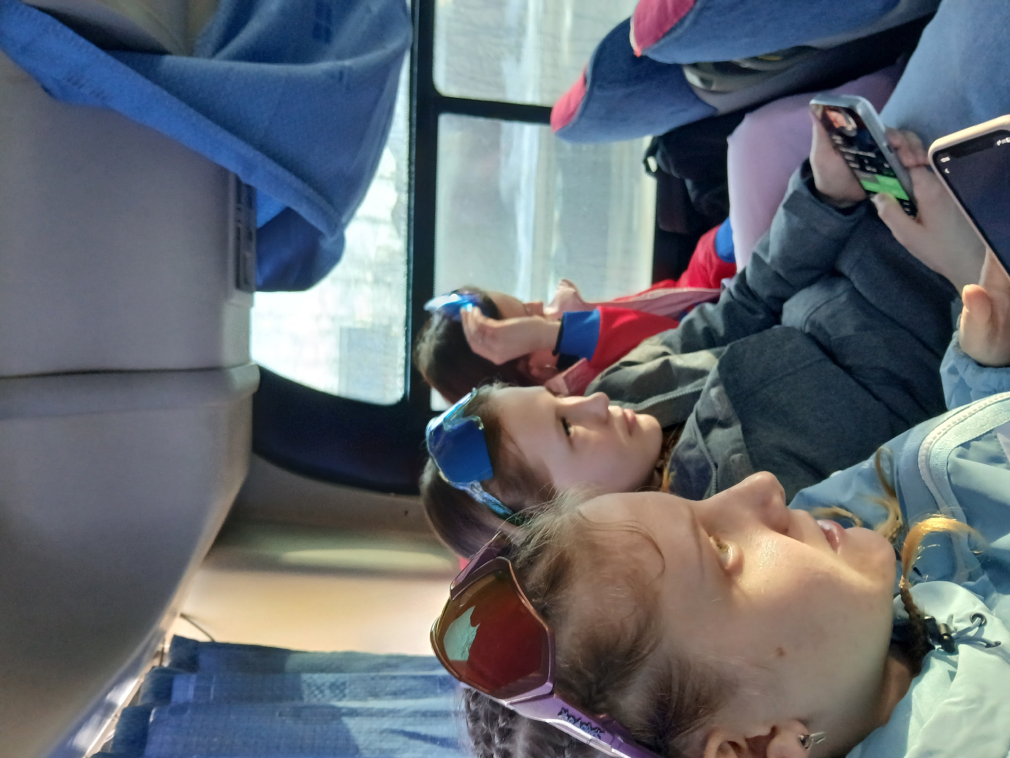 Three young people are lying down, possibly in a bus or other vehicle, with two of them using smartphones. The person closest to the viewer wears sunglasses perched on their forehead and a light blue jacket. The next person also wears sunglasses and a gray jacket, while the third person is mostly obscured but appears to wear a red and blue shirt. The interior suggests a travel setting. The location, Usolye, Russia, is not evident from the image itself.