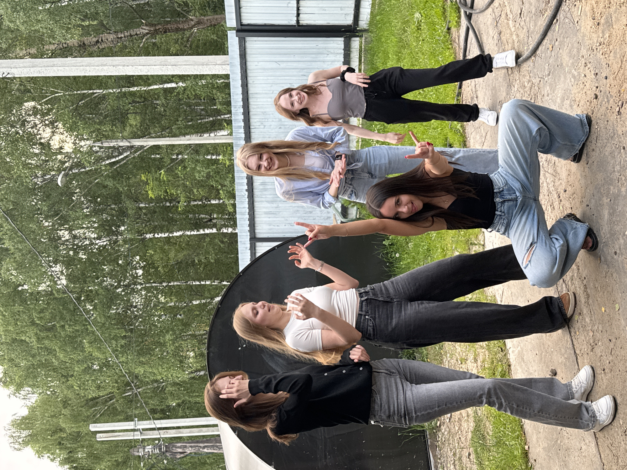 In a full-body, outdoor shot taken during the daytime in what appears to be a rural or suburban setting in Perm, Russia, five young women are posing for a photograph. The scene is outdoors with a weathered fence and lush green trees in the background. The ground is a mixture of dirt and concrete.
The women are arranged in a diagonal line, with the viewer's perspective being rotated 90 degrees clockwise. From bottom to top in the image, the first woman is wearing a black top and gray jeans, with her arm raised and hand covering her face. The second woman, also in gray jeans and a white t-shirt, has her hands up near her face and appears to be laughing or making a surprised expression. The third woman is dressed in ripped light-wash jeans and a black tank top, striking a peace sign with her fingers and smiling. The fourth woman is wearing a light blue button-down shirt over a white top and light-wash jeans, also smiling. The fifth woman at the top of the image is in black pants and a gray tank top, smiling and gesturing with her hands.
The overall mood of the photograph is joyful and energetic, with the women appearing to be having fun together. The lighting suggests it is daytime, possibly late afternoon, with bright but diffused light. There is no visible text in the image.