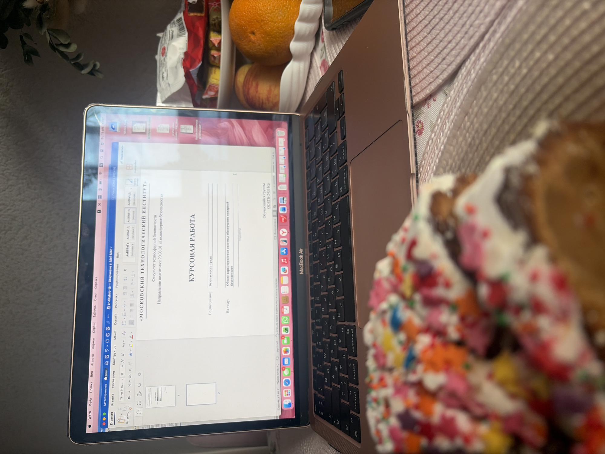 The image shows a laptop, likely a MacBook Air, open and displaying a Word document written in Russian. The document appears to be a course paper ("Курсовая работа") related to safety, specifically "Техносферная безопасность" (Technosphere safety), from the "Московский технологический институт" (Moscow Technological Institute). The laptop is placed on a surface, and next to it is food. The food includes oranges, an apple, and an Easter cake decorated with colorful sprinkles. The image suggests the person is working on their course paper while having snacks/food around them. The location cannot be determined based on the image.