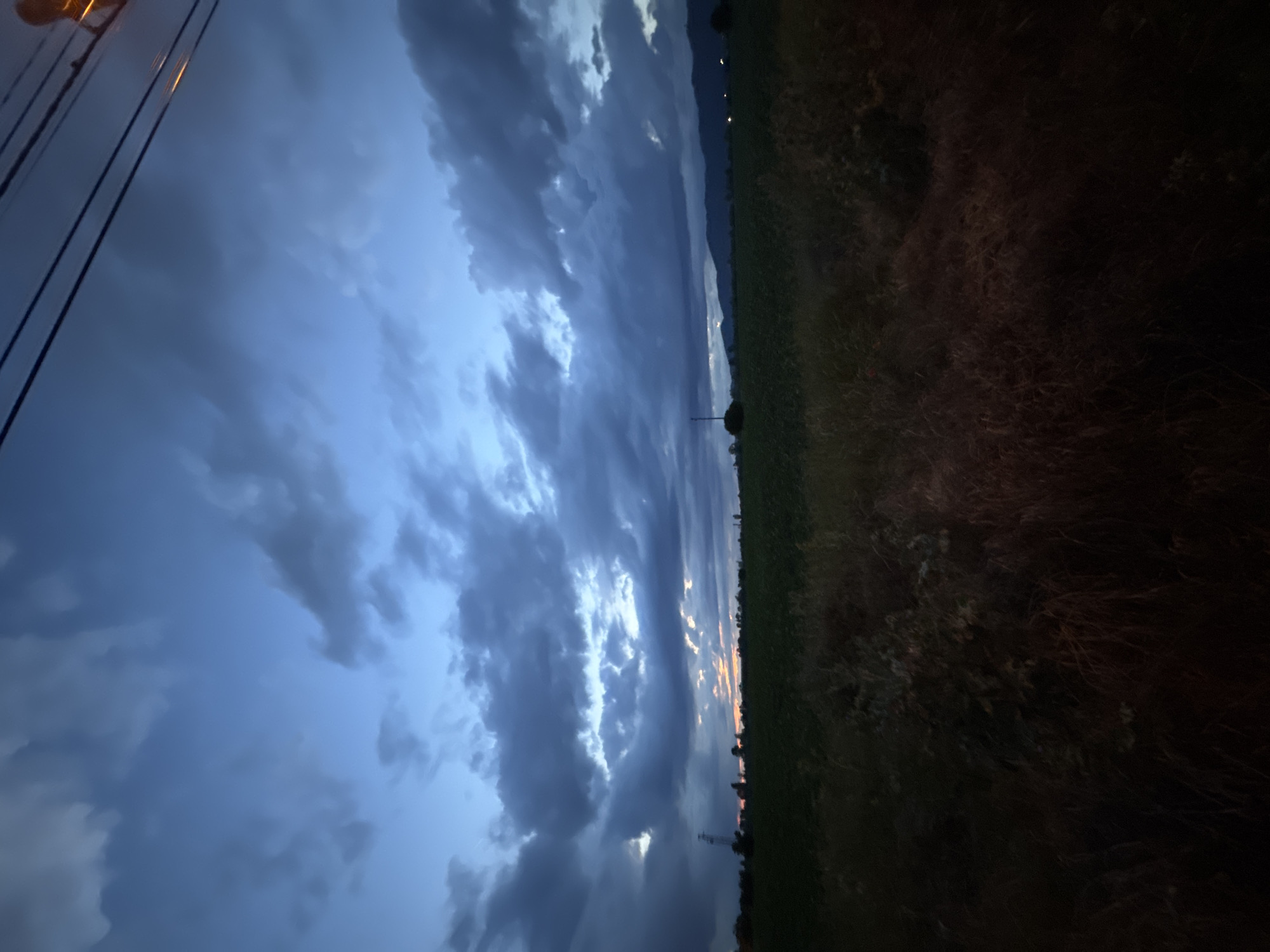 This landscape image, taken in Yana, Bulgaria, captures a dramatic twilight scene with an expansive, cloud-filled sky. The sky dominates approximately two-thirds of the frame, featuring heavy, dark blue-grey clouds that suggest an overcast evening or an approaching storm. Brighter patches of white and light blue are visible where the clouds thin, and subtle hints of orange and yellow light peek through along the distant horizon, indicating the recent setting of the sun.
On the far left, electrical power lines are visible, partially illuminated by a bright, warm orange-yellow light source, likely a streetlamp or a light from a building, situated just out of frame at the top. The wires lead to a utility pole, the top of which is faintly discernible.
The right side of the image depicts a dark field of grass and unkempt vegetation, rendered in deep greens and browns due to the low light. In the middle ground, a lone, small tree or bush stands silhouetted against the sky. Further in the distance, a dark, undulating line suggests mountains or hills that form the backdrop. Along the base of these distant features, several faint, scattered lights can be seen, possibly indicating a remote village or isolated buildings.
The scene conveys a sense of peacefulness and vastness, characteristic of a rural or semi-rural setting at the transition from dusk to night. There is no discernible activity or any individuals present in the image. No text is visible.