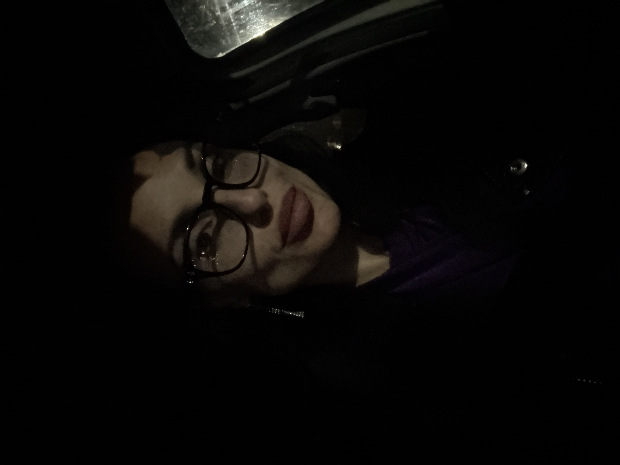 A woman with glasses is lying in what appears to be a car. The lighting is very dark, but it is possible to discern the outline of her face, glasses, and lips, which are visible. A part of a car's interior, perhaps the roof, can be seen above her. There are no other identifiable features, and no information about the location is available to place the car in Pleven, Bulgaria.
