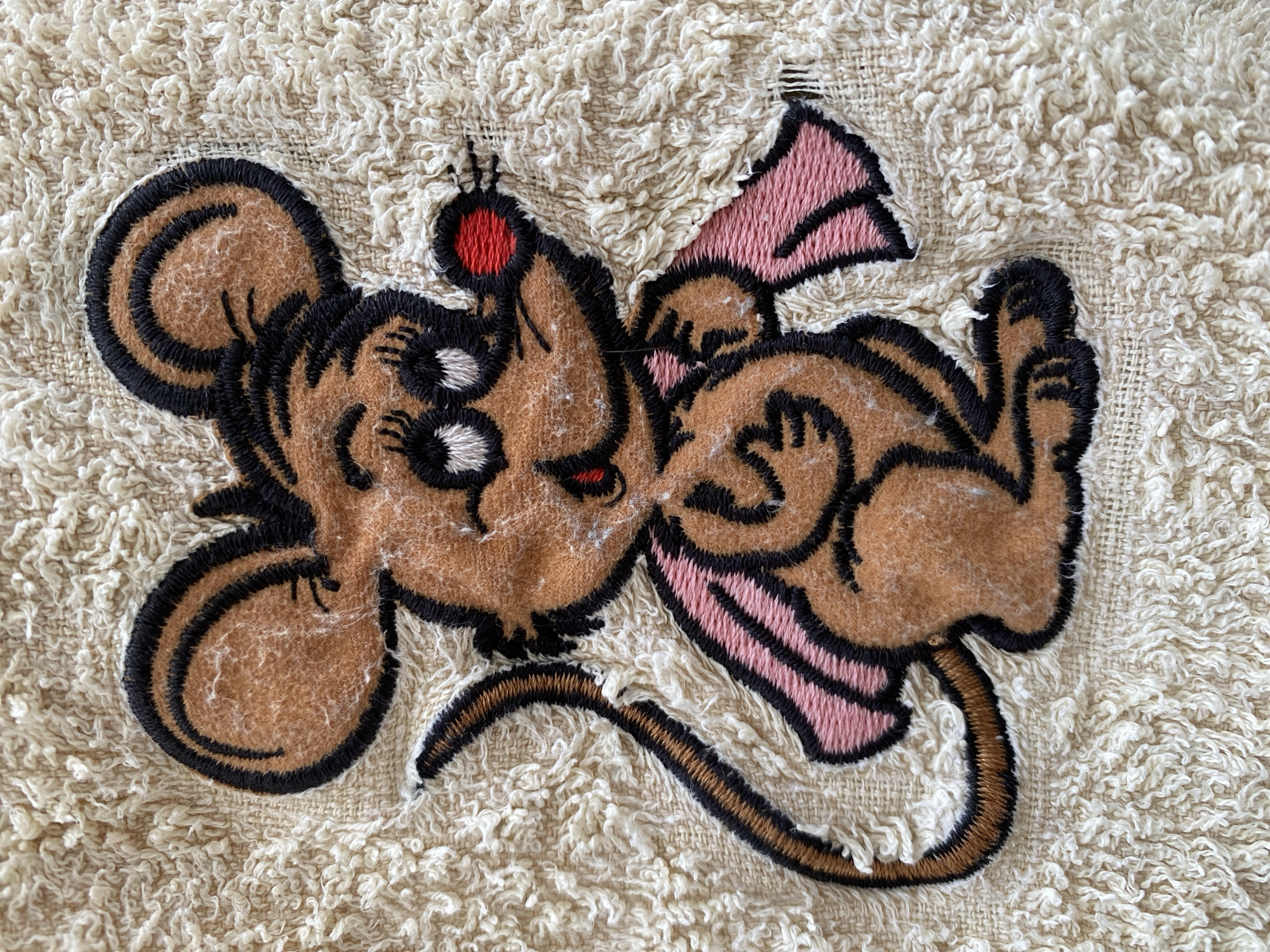 An embroidered patch featuring a brown cartoon mouse is centrally displayed against a cream-colored, looped fabric background, likely terrycloth. The mouse is outlined in black stitching and possesses large, round black eyes with white pupils and eyelashes, a circular red nose, and a small red smile. Its large, round ears are brown with black outlines, matching the body. A thin, brown tail with a black outline extends upwards from the left behind the mouse. A pink scarf or shawl, also outlined in black, is draped around its neck and shoulders, with two ends extending laterally. The brown and pink areas of the patch exhibit a slightly fuzzy texture. The background fabric shows a dense, irregular loop pile with minor wear and loose fibers visible, especially around the edges of the patch stitching. This detailed image of the patch and its fabric background was captured in Budva, Montenegro.