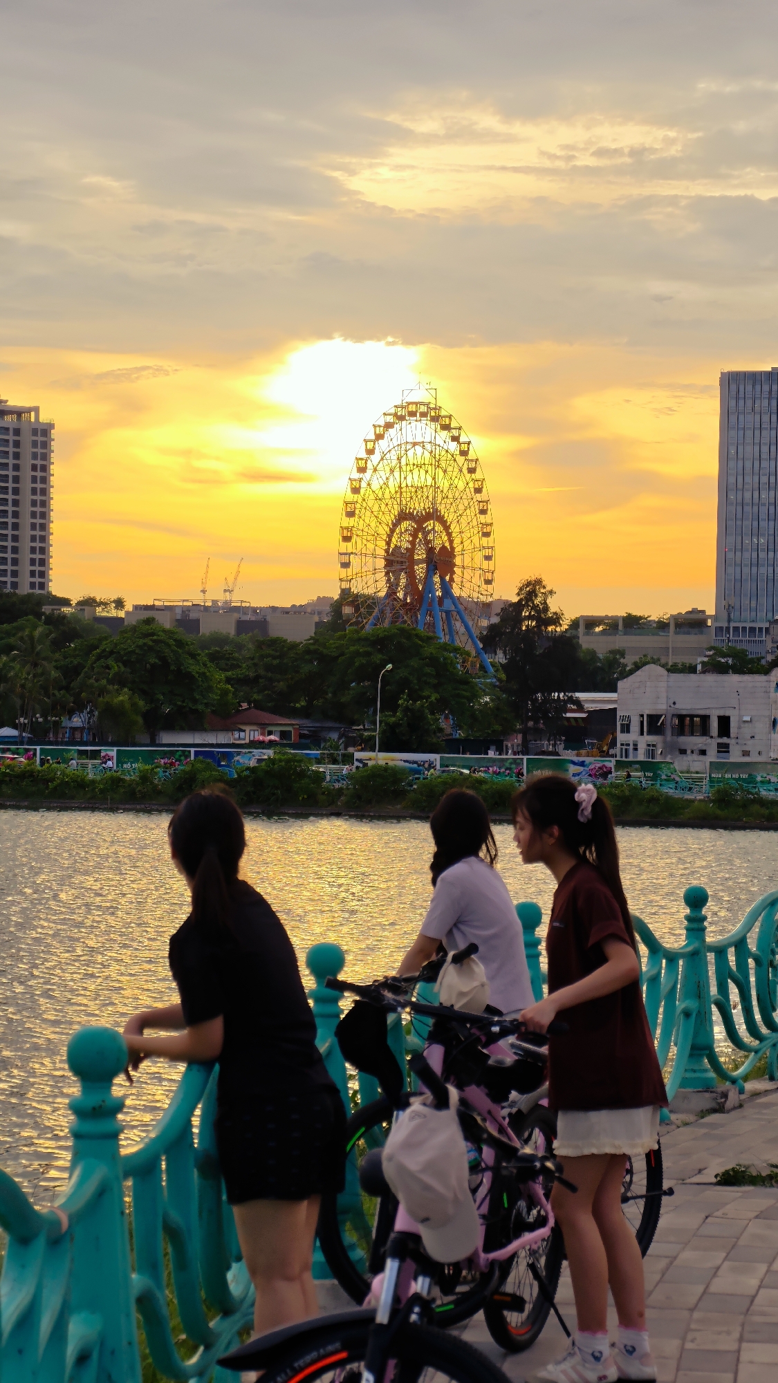This media file captures a scene at dusk along a waterfront in Hà Nội, Vietnam. Three young women are present, each with a bicycle, suggesting they are enjoying an evening ride or outing. The woman on the left, seen from behind, wears a black dress and leans on a turquoise railing. In the center, a woman in a white t-shirt is positioned between two pink bicycles, one of which has a beige cap hanging from its handlebars. To the right, a woman in a maroon t-shirt and a white skirt stands with her bicycle, wearing white socks and sneakers.
The background is dominated by the vibrant glow of the setting sun, casting a warm, golden-orange hue across the sky and reflecting on the water. A large Ferris wheel, illuminated and prominent, stands out against the sky, indicating the presence of an amusement park or entertainment area. Tall buildings, including a modern high-rise on the right and another on the far left, suggest an urban setting. Lush green trees line the waterfront, separating the path from the distant buildings and Ferris wheel.
The overall mood appears serene and picturesque, with the warm lighting of the sunset creating a peaceful atmosphere. The scene is set by a river or lake, with a decorative turquoise railing running along the edge of the walkway. The time of day is clearly sunset due to the intense colors in the sky and the warm light. There is no visible text within the image.
