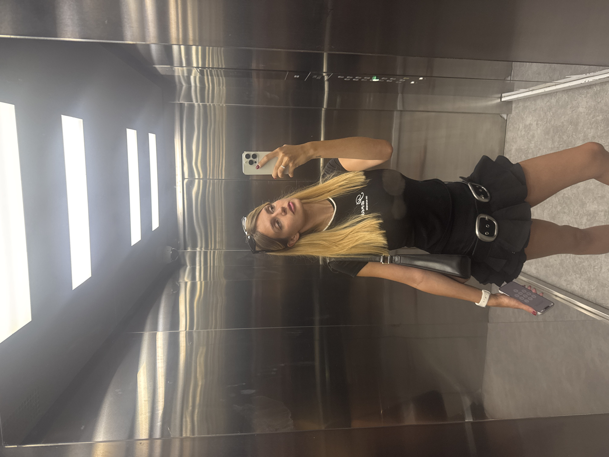In an elevator with stainless steel walls and fluorescent lighting, a woman with blonde hair styled down takes a mirror selfie. She is wearing a black t-shirt, a black pleated skirt with a prominent belt, and a white smartwatch. She holds a smartphone in her right hand, and a black handbag is slung over her left shoulder. The elevator's control panel, featuring illuminated buttons, is visible on the right side of the frame.