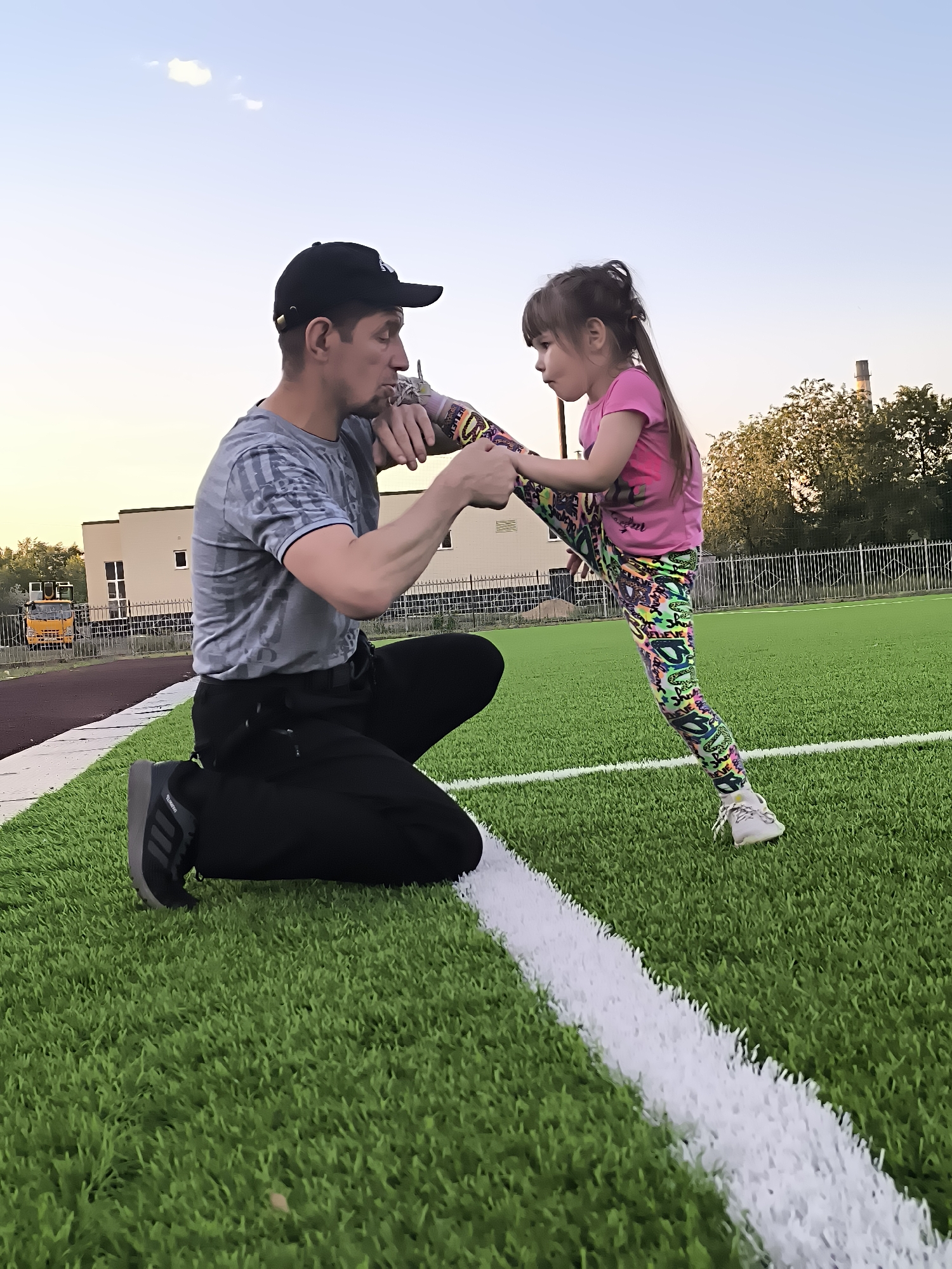 An adult male wearing a grey t-shirt, black pants, and a black baseball cap is kneeling on artificial turf, assisting a young girl. The girl, dressed in a pink t-shirt and colorful patterned leggings, is standing with one leg extended. They are on a sports field with a white line marking the edge of the field in the foreground. In the background, there are buildings, trees, a fence, and a bright sky. The scene appears to be taking place during daylight hours.