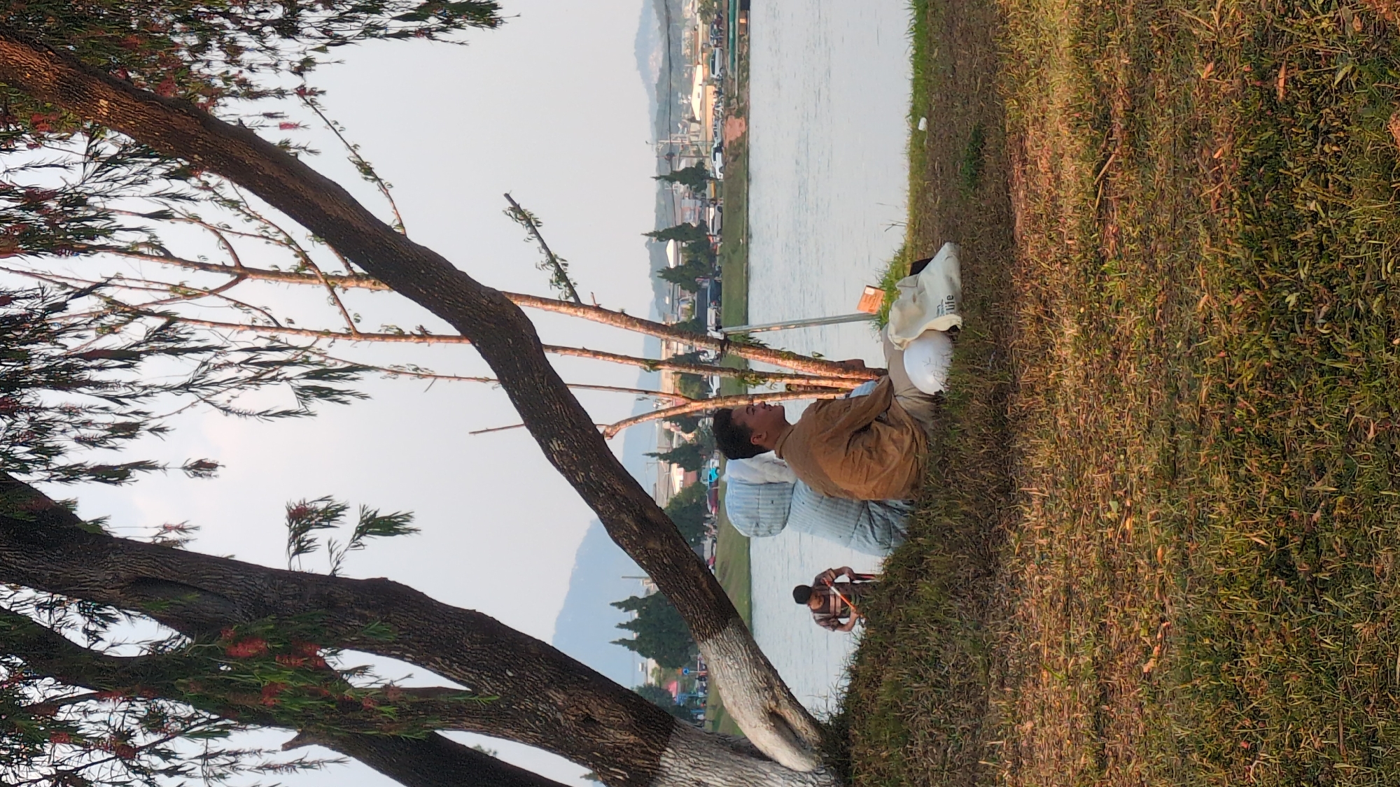 Captured in the city of Da Lat, Vietnam, the scene portrays individuals alongside a body of water. Three people are visible; two are seated near a tree trunk and one is on the ground and appears to be photographing or filming near the water. A white bag and a white hat are placed beside the two people who are seated. In the background, buildings and vegetation line the opposite shore of the water. A large tree frames the left side of the image, and its branches extend into the scene. Grass covers the ground in the foreground. The overall lighting suggests it is daytime.