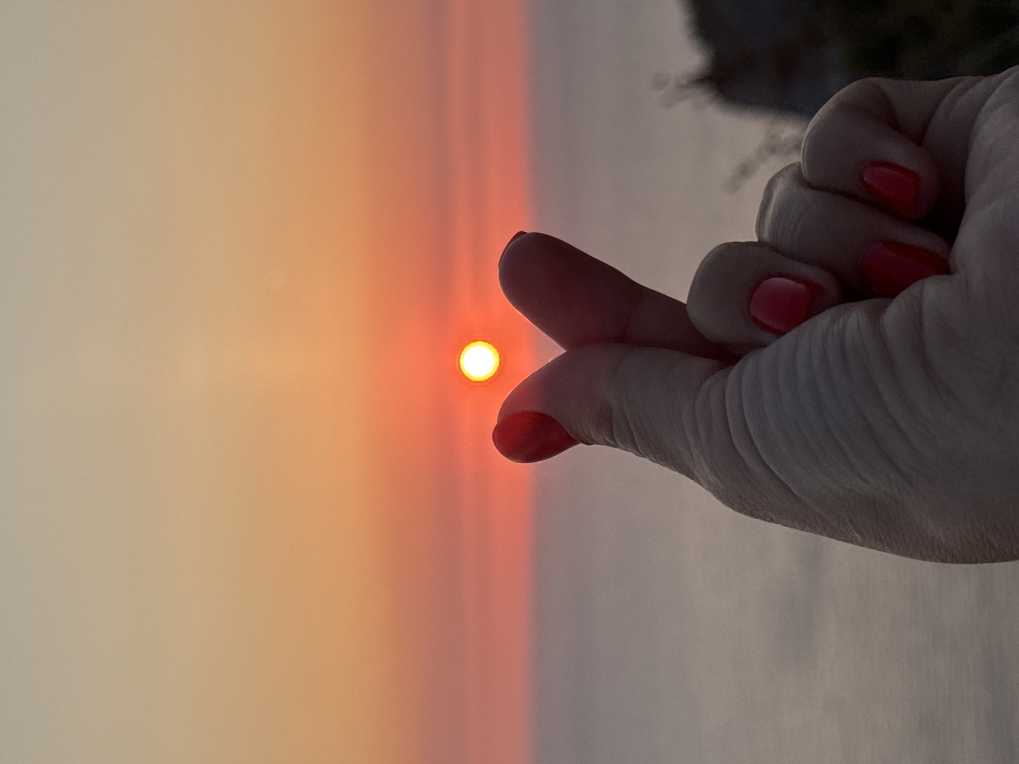 An extreme close-up shot focuses on a person's hand making a finger heart gesture in front of a sunset over the sea. The hand, painted with bright red nail polish, is silhouetted against the vibrant orange and pink sky. The sun, a small, glowing orb, is positioned between the thumb and index finger of the hand, creating a sense of capturing or framing the sunset. The sea is visible in the background, stretching to the horizon, and a faint outline of a cliff or hillside can be seen on the right. The lighting suggests it is either sunrise or sunset. The image was captured in Donji Katun, Montenegro.