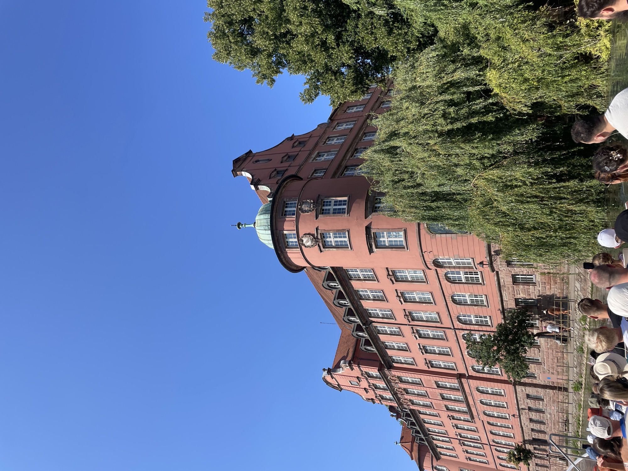 The image shows a large, salmon-pink building with a red tiled roof and numerous white-paned windows, situated along a canal. A round tower with a green domed roof is visible on the right side of the building. Lush green trees, including a weeping willow, partially obscure the building's facade. In the foreground, the backs of several people are visible, suggesting they are on a boat. The sky is a clear, bright blue. This building is located in the Petite France quarter of Strasbourg, France, a historic area known for its picturesque canals and traditional Alsatian architecture.